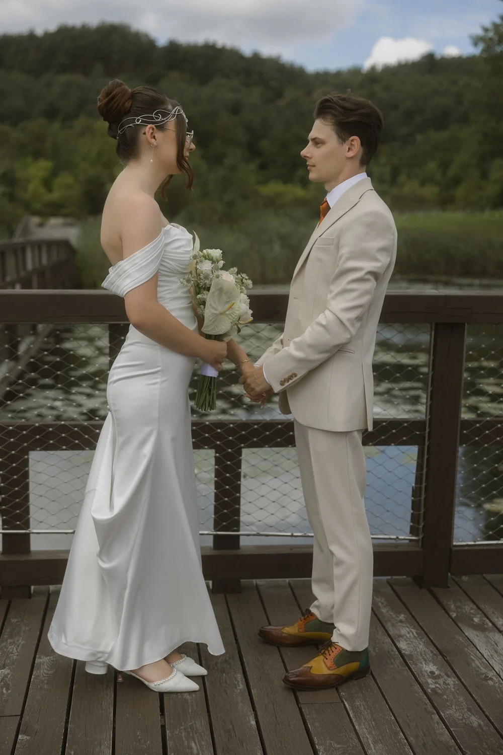 A bride and groom holding hands on a wooden bridge, with a river and forested landscape in the background, during their wedding ceremony.