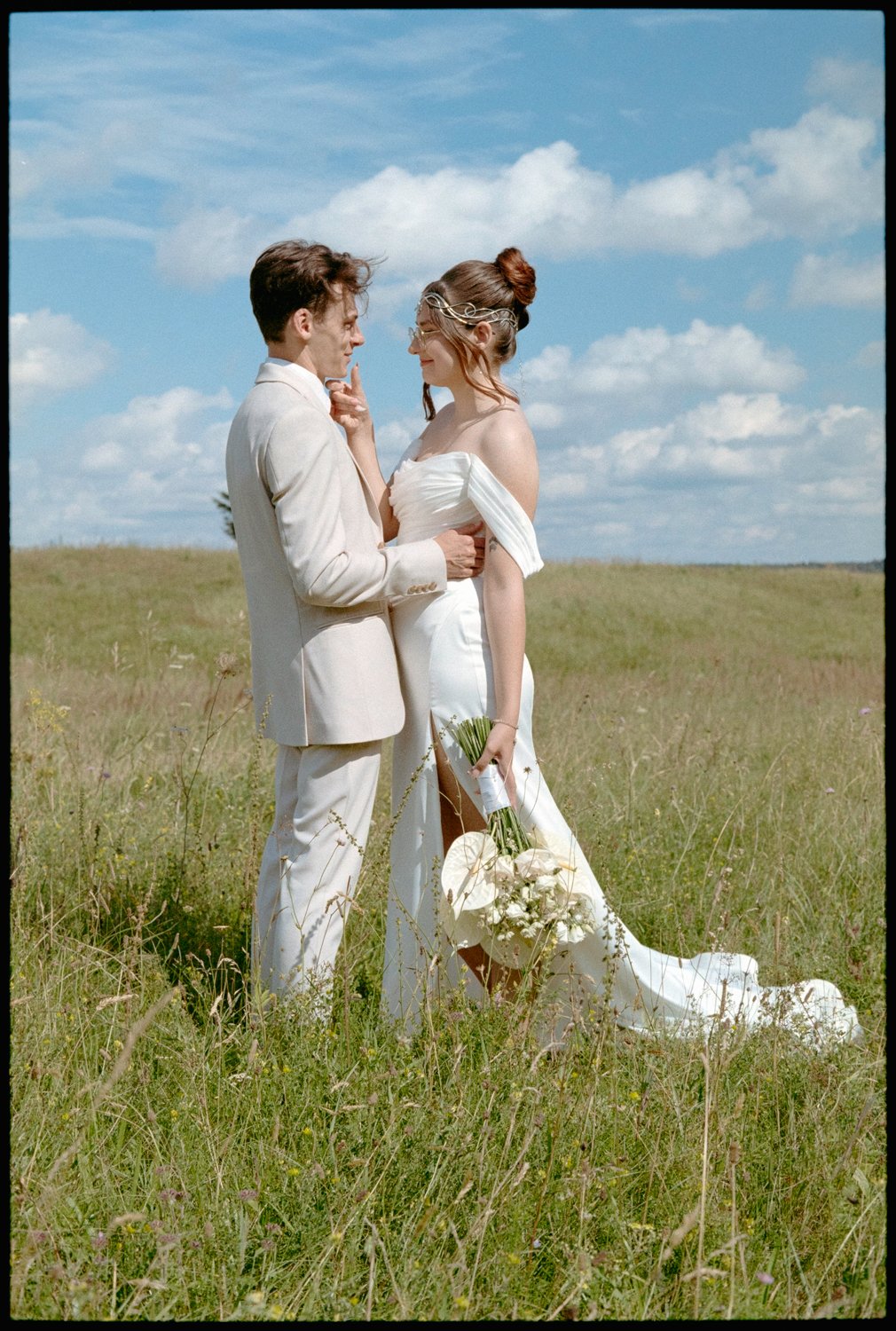 A couple in wedding attire standing in a grassy field under a blue sky with clouds, holding a bouquet of white flowers.