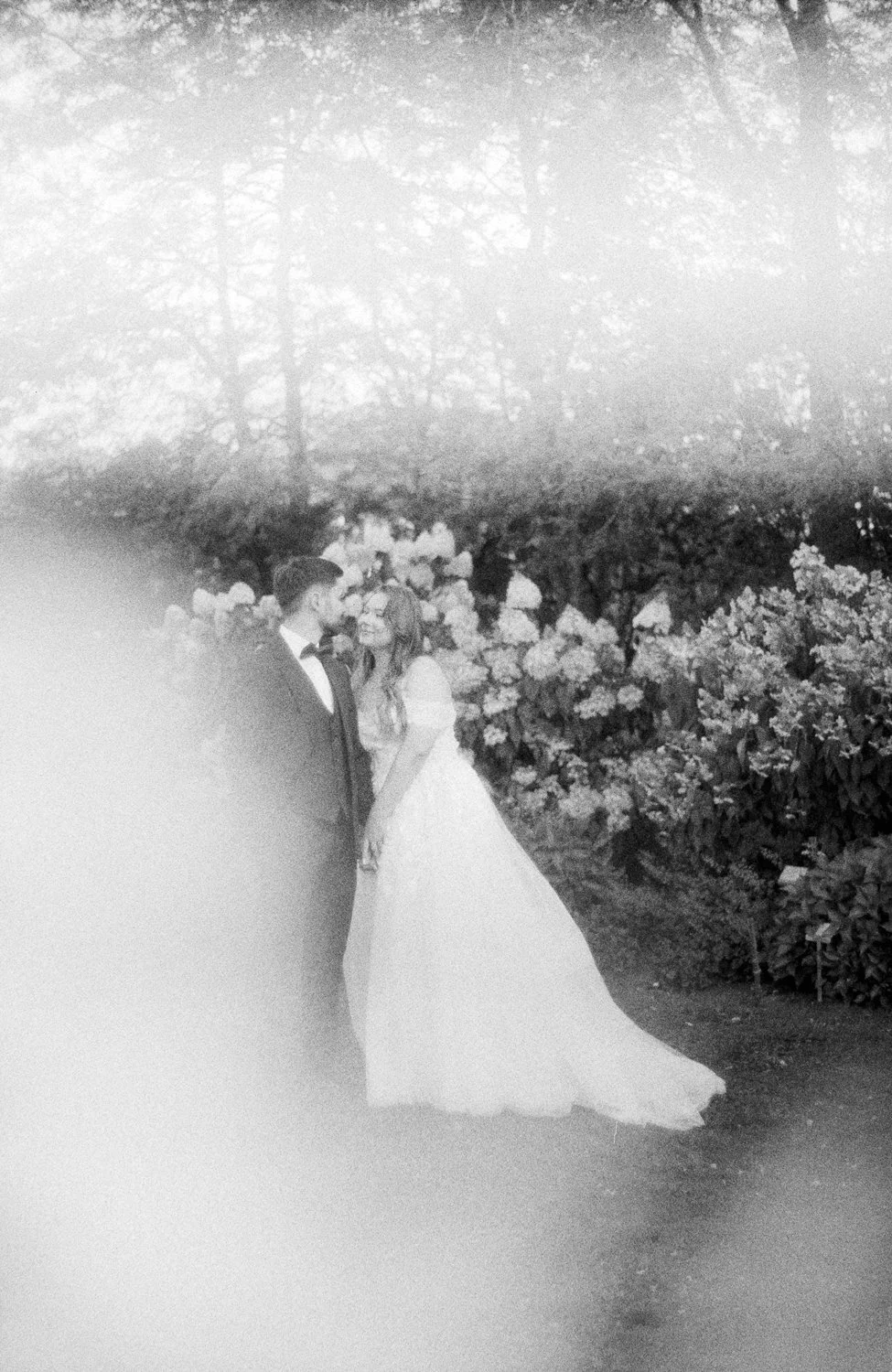 A black and white photo of a bride and groom standing closely and holding hands in an outdoor garden with trees and blooming flowers.
