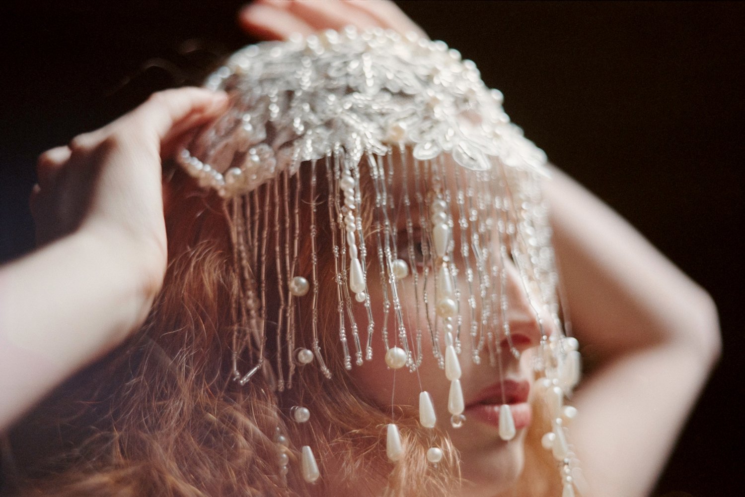 A person with long red hair is wearing a large, ornate, pearl and beaded headpiece that covers their face, with the person's hands adjusting or holding the headpiece.