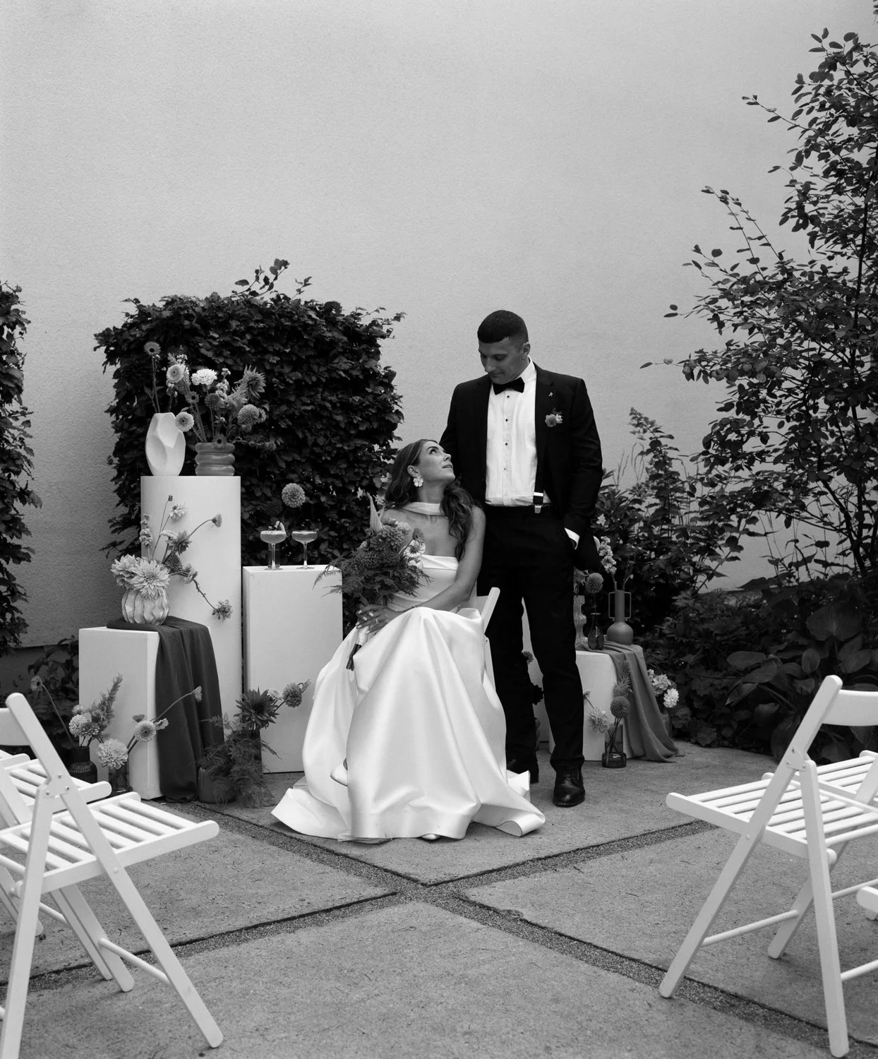 A bride and groom at their wedding, with the bride seated in a white dress holding a bouquet, and the groom standing beside her in a tuxedo, looking down at her. There's a decorated outdoor setting with flowers, vases, and chairs around them.