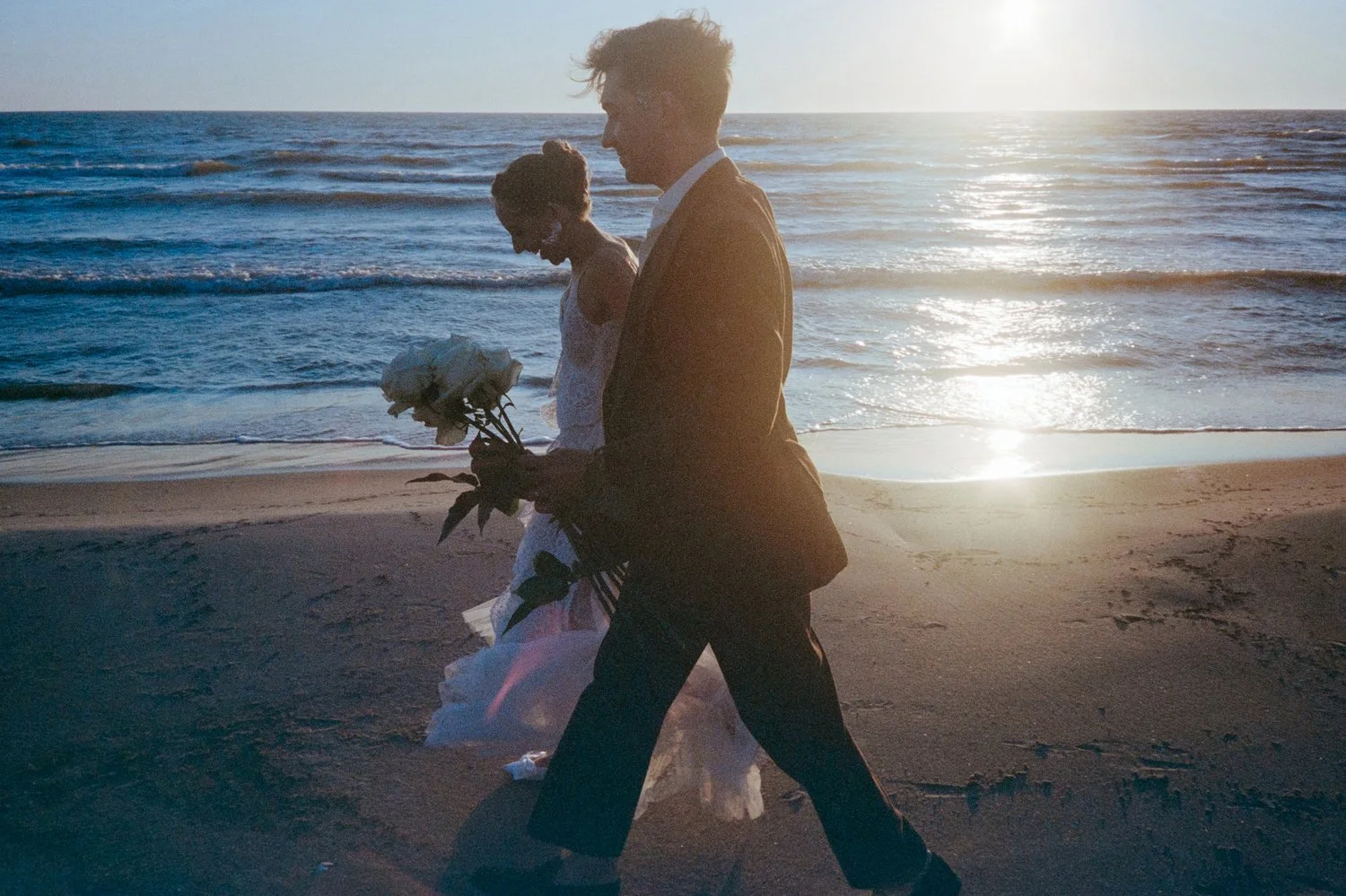 A man in a suit and a woman in a white dress walking along the beach at sunset, with the man carrying a bouquet of white flowers.