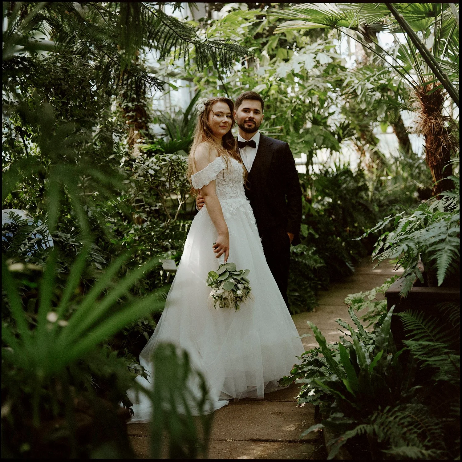A bride and groom posing together in a lush greenhouse or botanical garden surrounded by green plants and foliage.