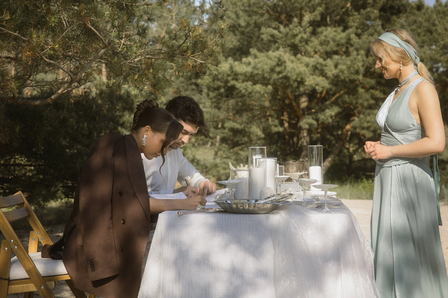 A couple signs marriage license at an outdoor wedding table with a woman standing nearby. The table has white candles and silverware, surrounded by trees.
