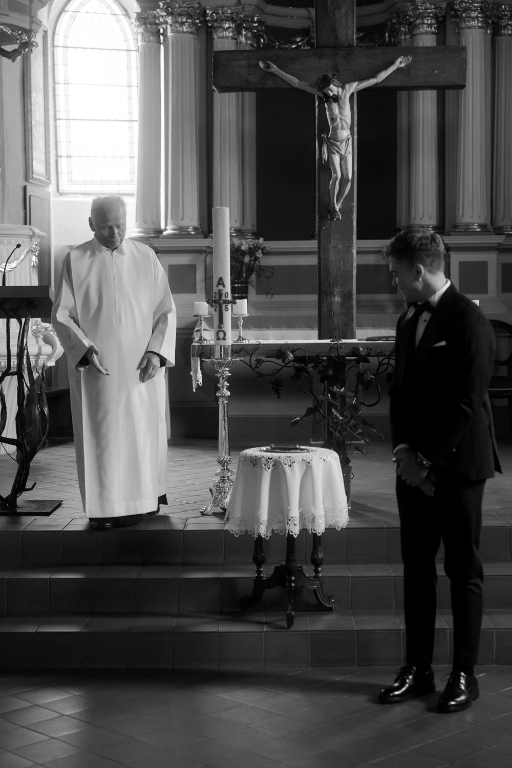 A priest and a young man in formal attire praying in a church during a religious ceremony, with a crucifix hanging in the background.