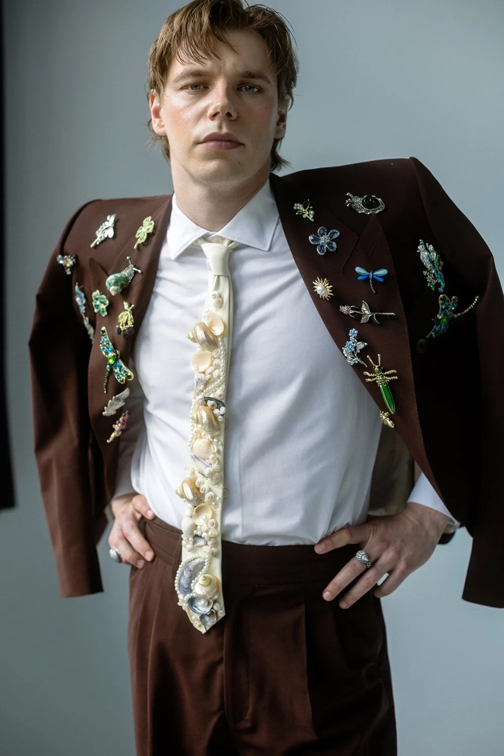 A man wearing a brown suit jacket adorned with various colorful brooches, a white shirt, and a tie decorated with seashells and pearls. He has his hands on his hips and looks directly at the camera.