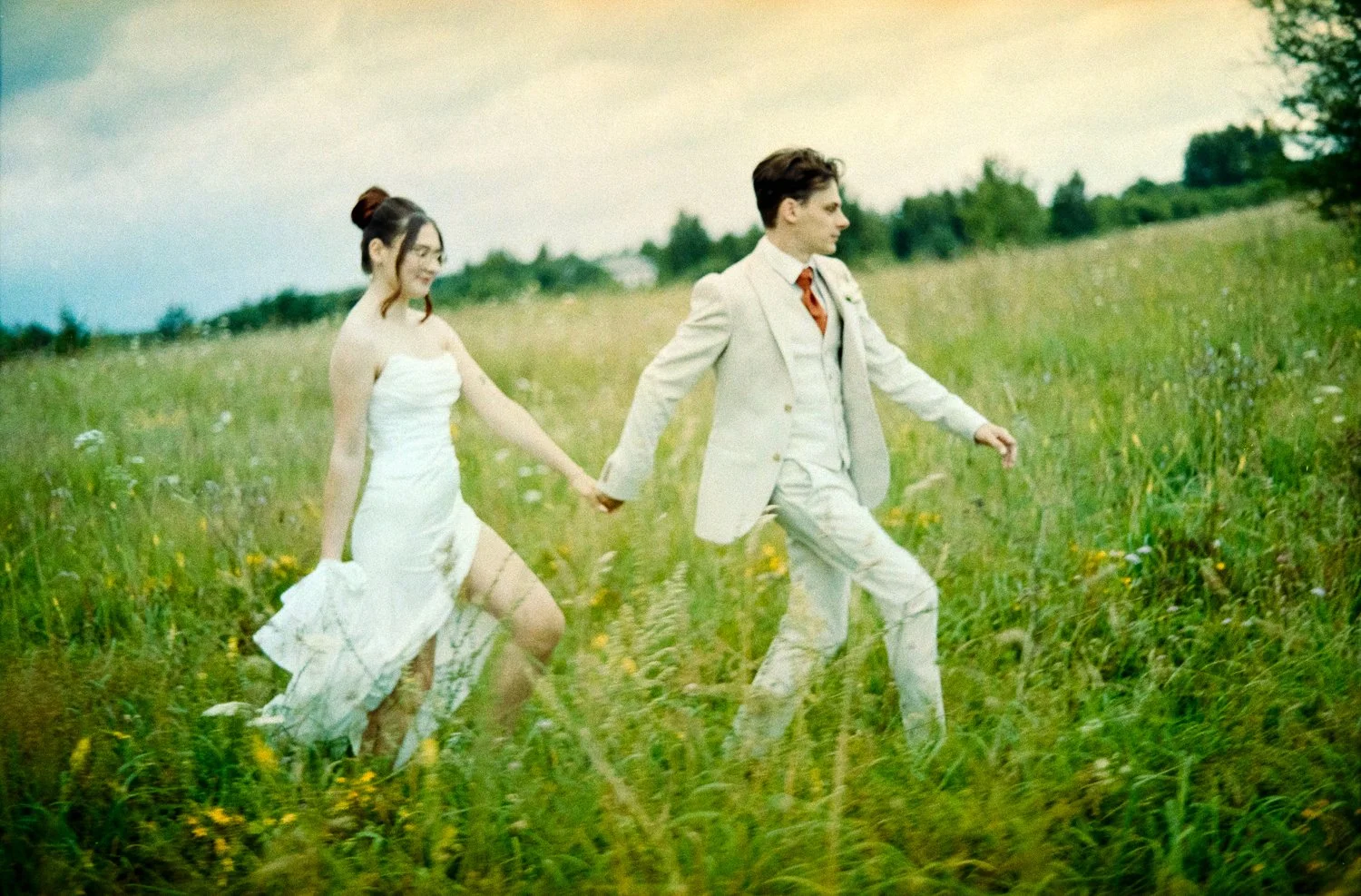 A couple dressed in wedding attire walking hand in hand through a grassy field.