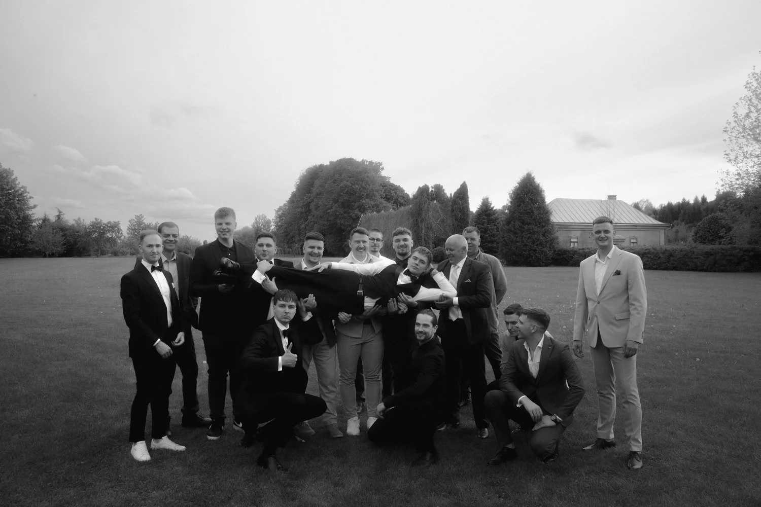 A group of men in formal suits outdoors holding a young man horizontally in their arms, with a large open field, trees, and a building in the background, in black and white.
