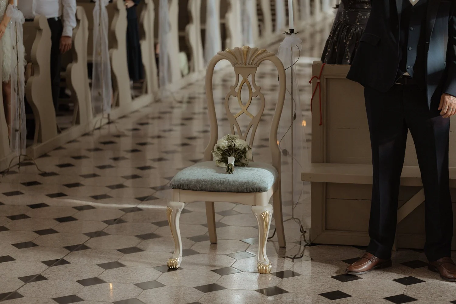 Wedding ceremony with a bouquet on an ornate chair, in a decorated church with guests standing along the aisle.