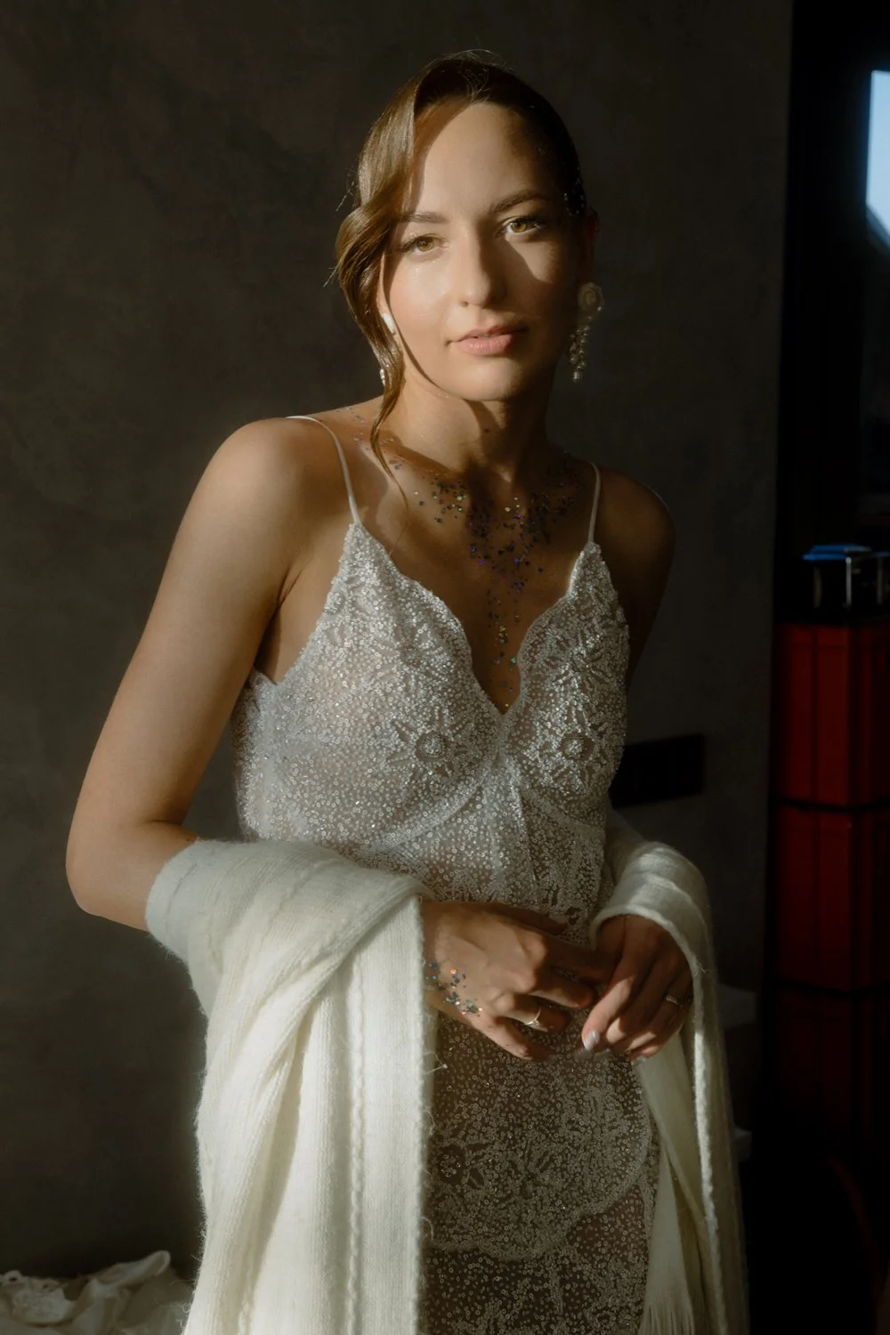 A woman with light brown hair styled in waves, wearing a delicate white lace dress with spaghetti straps, jewelry, and a white shawl.