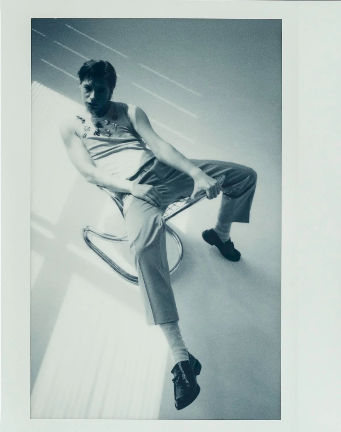 Black and white photo of a young man with short hair sitting on a modern metal chair, with one leg crossed over the other, wearing a sleeveless top, loose trousers, and sneakers, in a room with horizontal blinds casting light patterns.