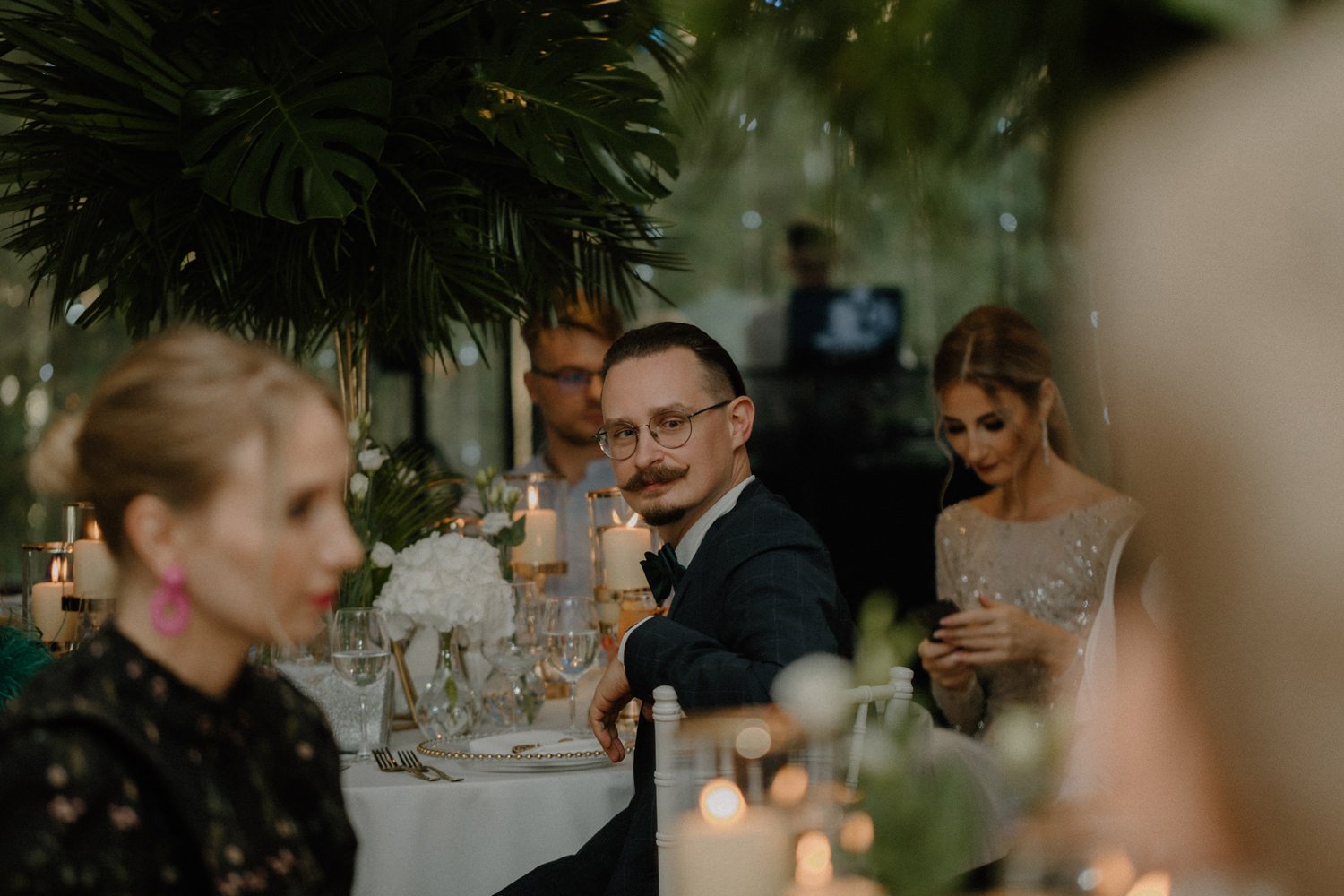A man with glasses and a mustache in a tuxedo sitting at a decorated dining table during an event, surrounded by others, with candles and floral arrangements, in an elegant indoor setting.