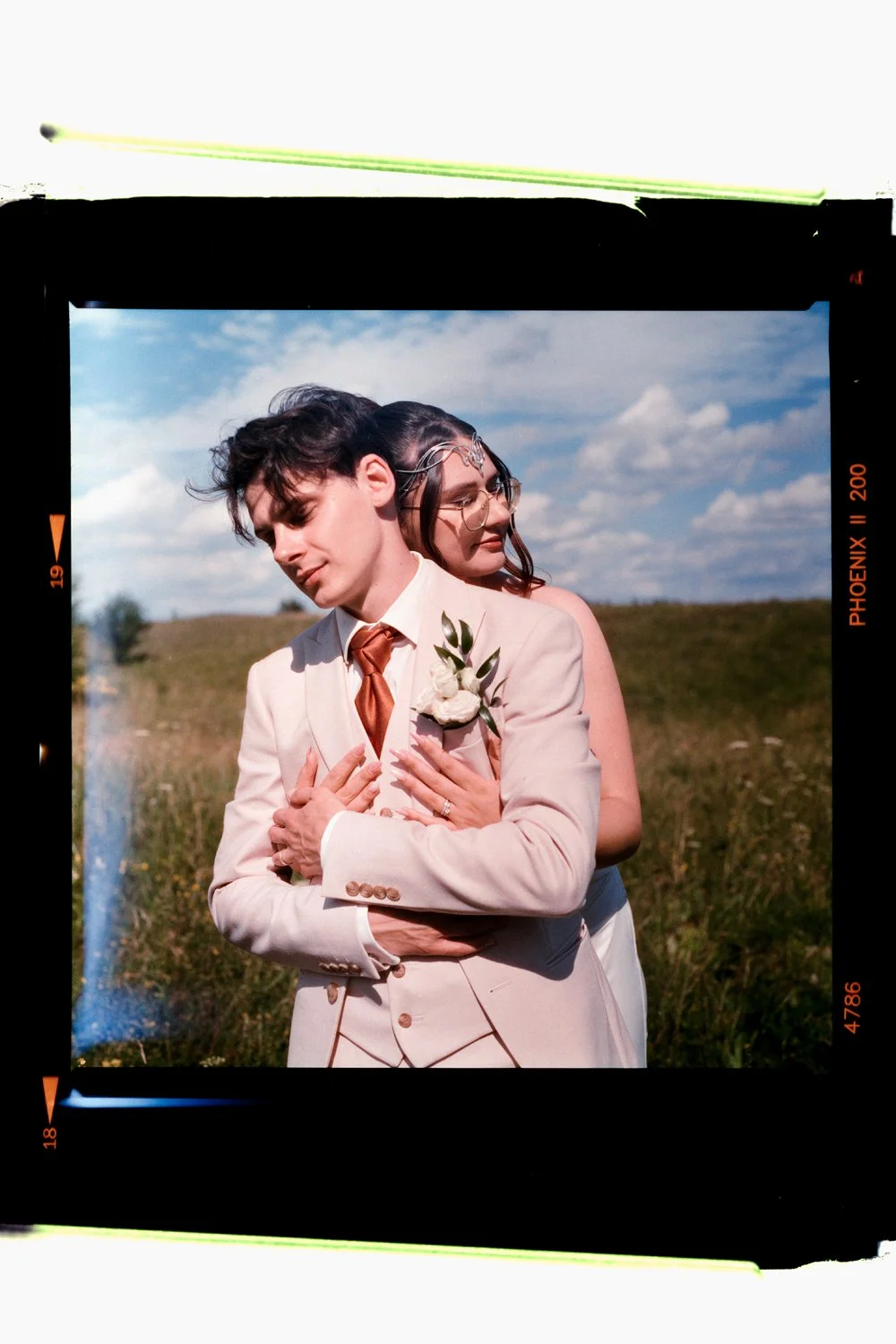 A groom in a beige suit with a boutonnière and a bride in a wedding dress embrace outdoors against a landscape with grass and a blue sky with clouds.