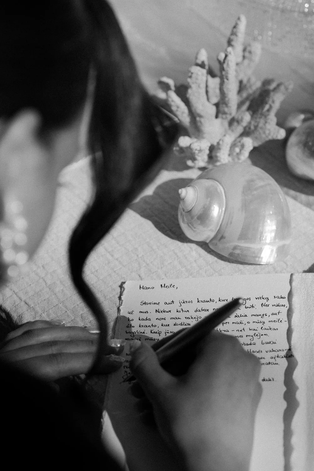 A person writing a letter with a pen, with seashells and a coral piece on the table, viewed from above in black and white.