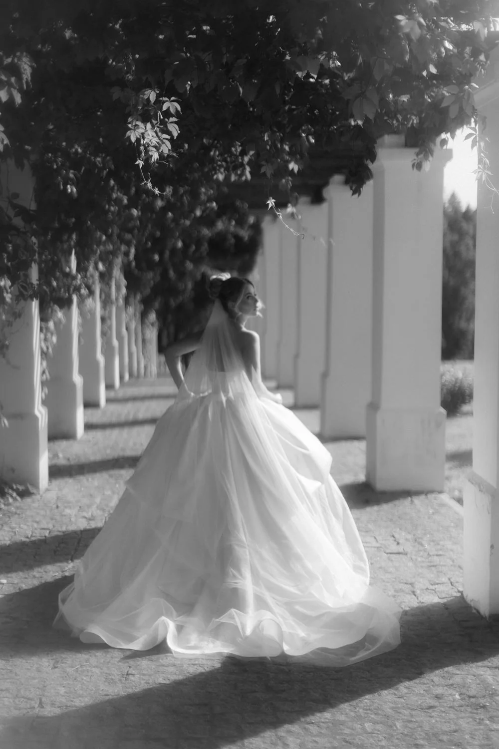 A woman in a wedding dress standing under a canopy of tree branches, looking to the side in a garden or outdoor setting.