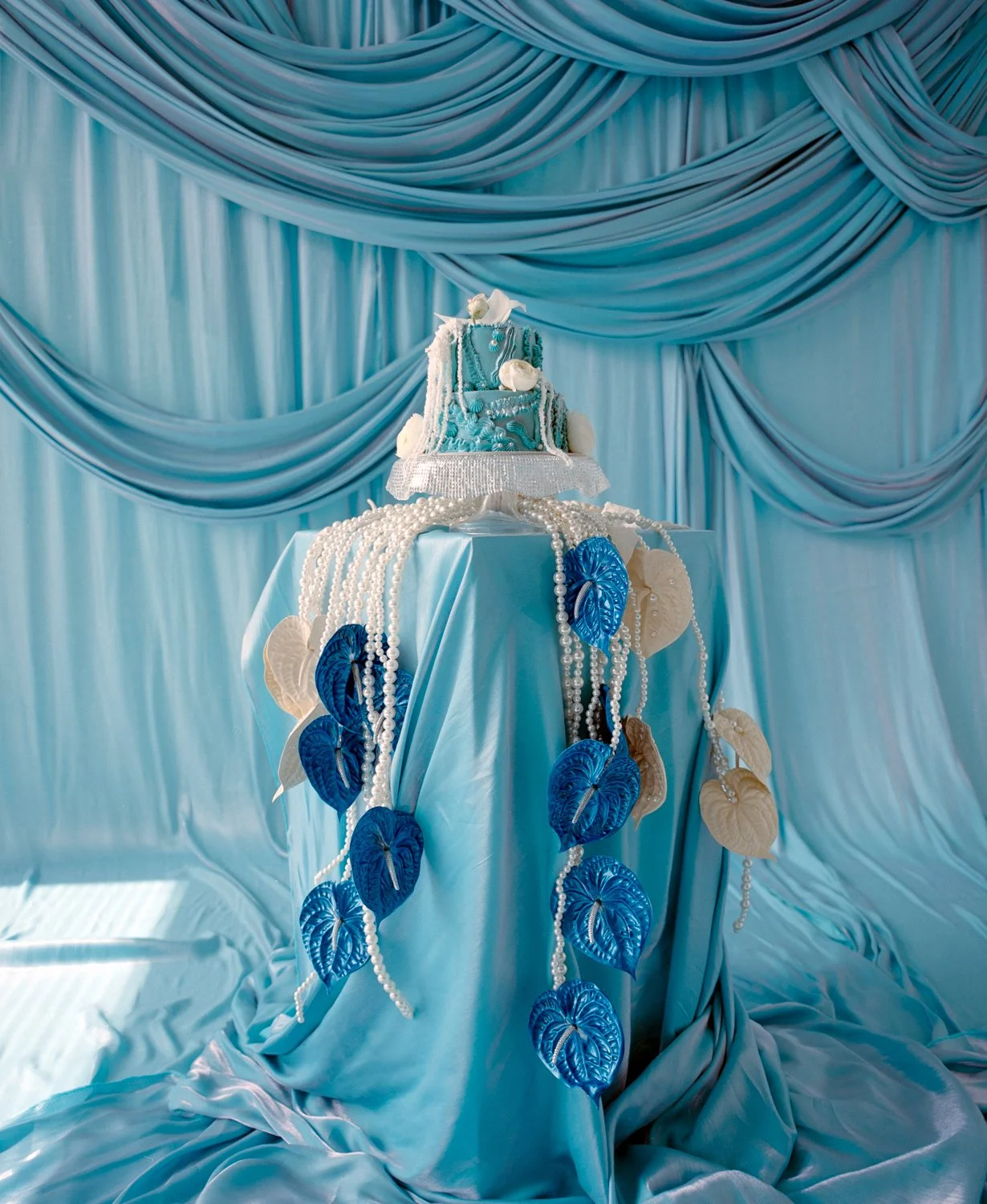 Blue draped backdrop with a tiered cake decorated with white flowers and blue accents, displayed on a table covered with a blue satin cloth and adorned with strings of white pearls and blue and white leaves.