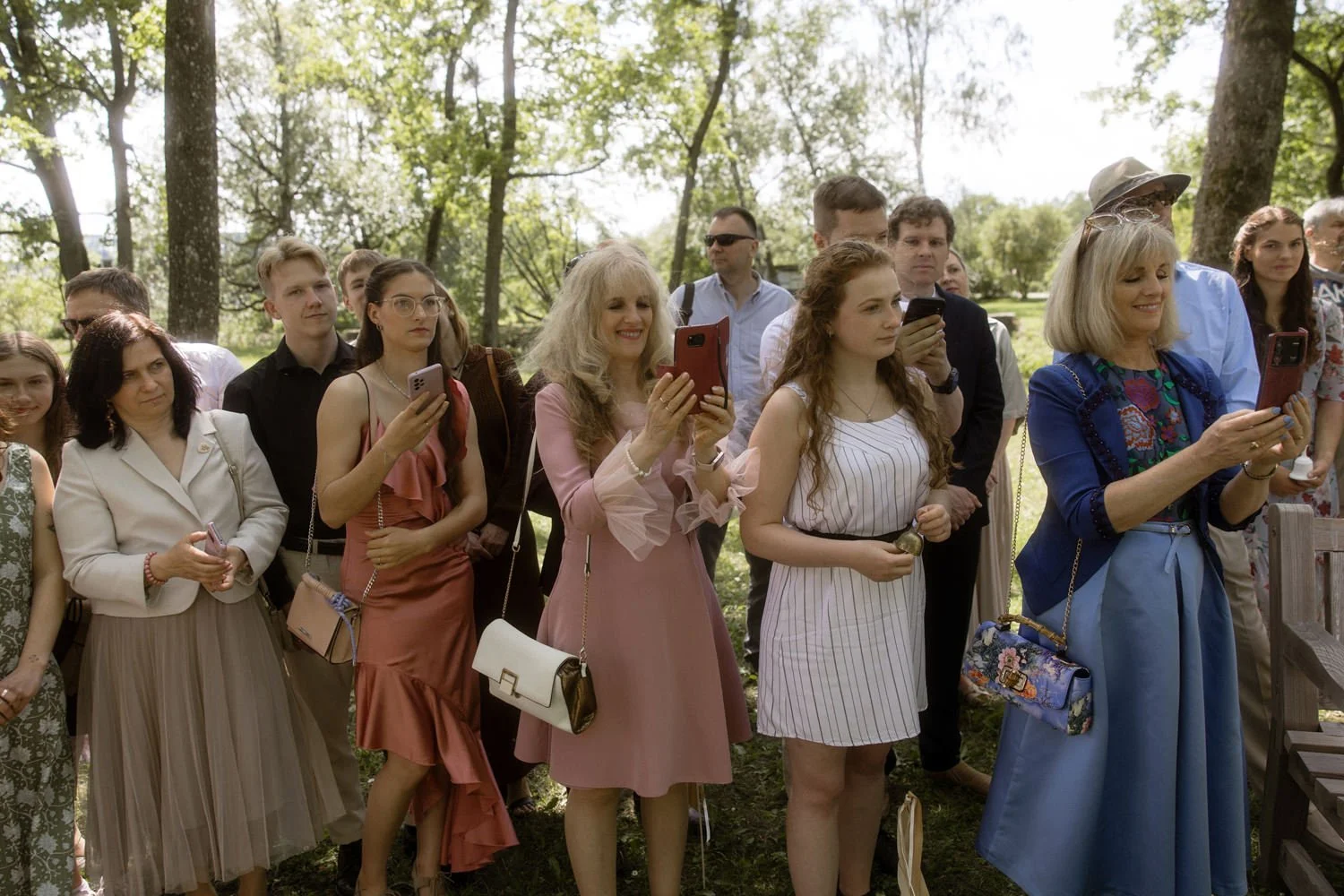 People standing outdoors in a park, many looking at their phones, dressed in semi-formal attire.