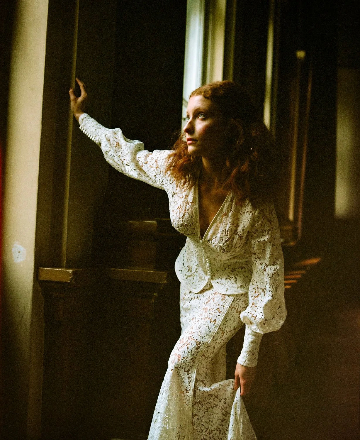 A woman with curly red hair dressed in a white lace suit, standing indoors, looking toward a window with soft natural light illuminating her face, in a vintage or classic setting.