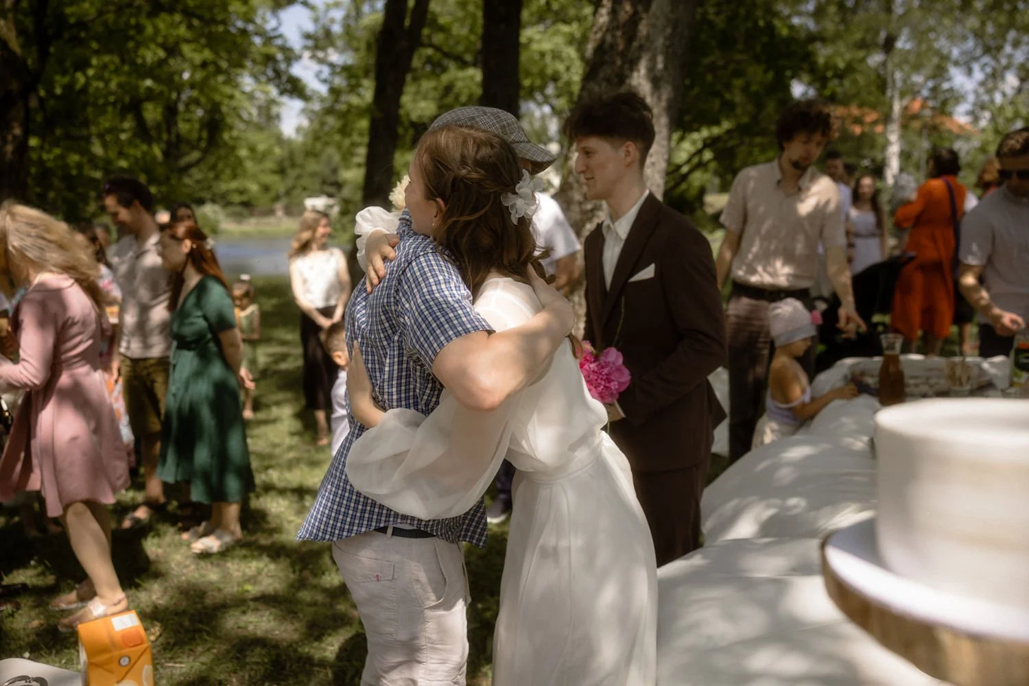 People embracing at an outdoor wedding reception with trees and a lake in the background.