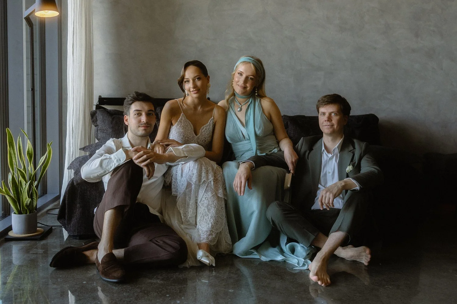 Four people sitting on a couch in a living room, two men and two women, dressed for a formal occasion, with a plant and a window with curtains nearby.