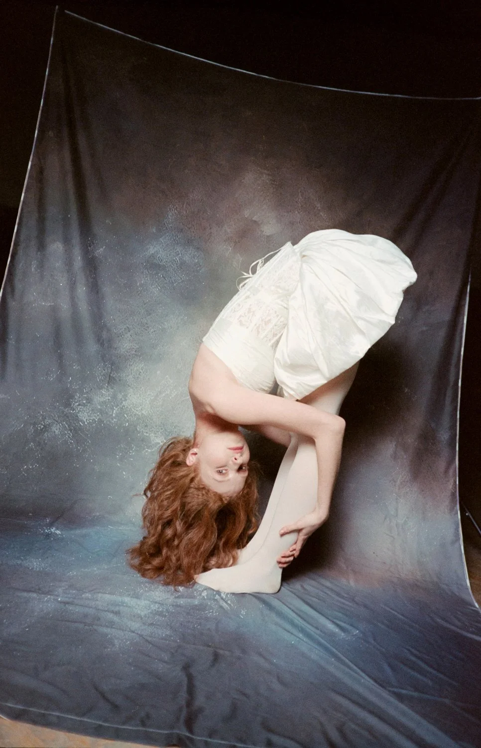 A woman with long, curly red hair is posing in a curled-up position on a black backdrop, wearing a cream-colored dress, with her head turned upside down facing the camera.
