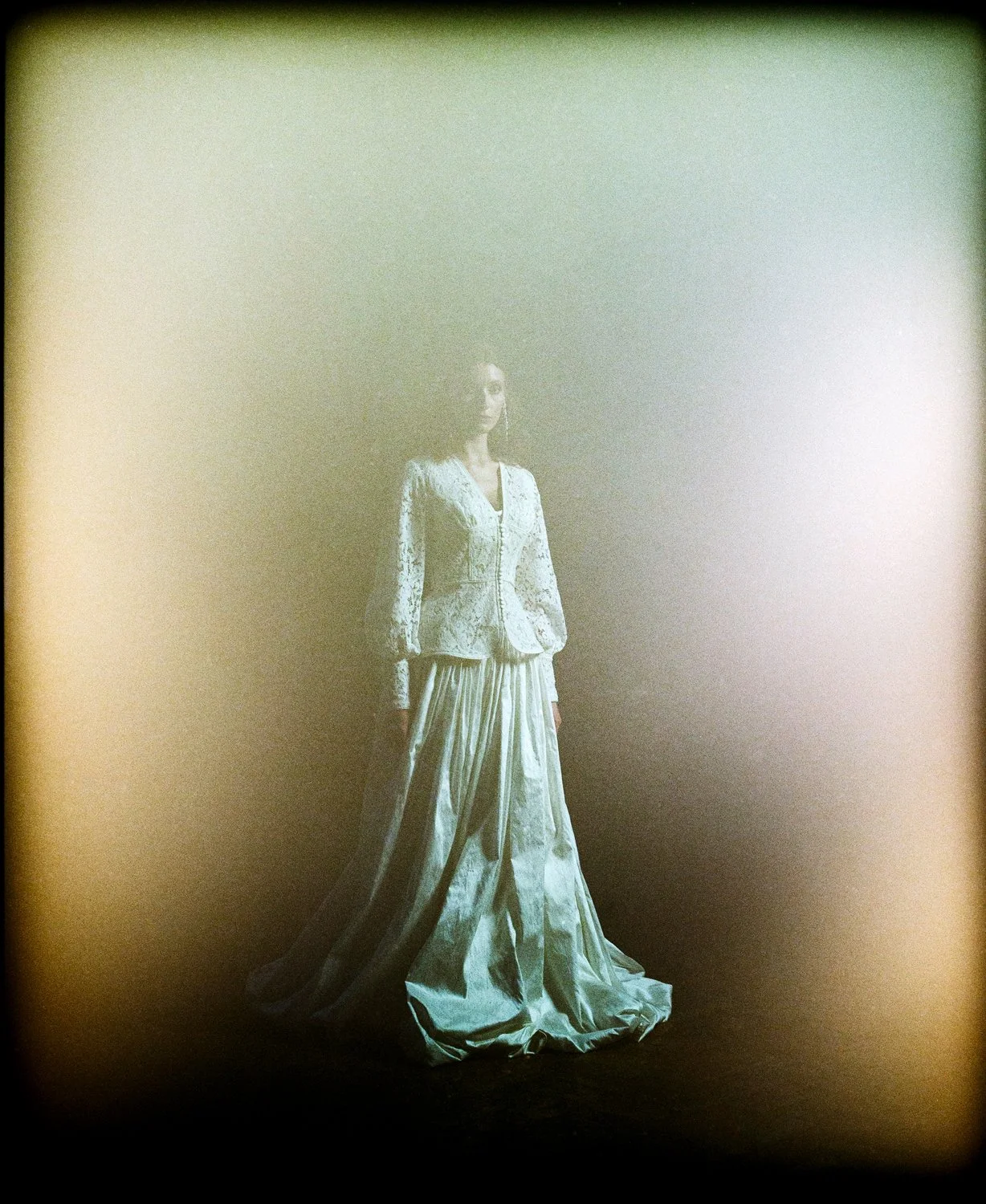 A woman wearing a vintage white lace suit and wide-leg pants standing alone against a dark background, with a hazy light effect around her.