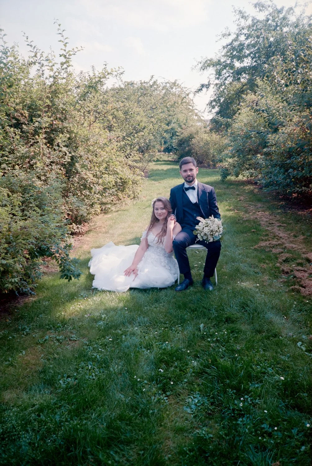 Bride and groom sitting together outdoors in a lush, green garden during daytime. The bride is in a white wedding dress, sitting on the grass, while the groom, in a navy suit with a bow tie, is seated on a chair holding a bouquet of white flowers. Bo