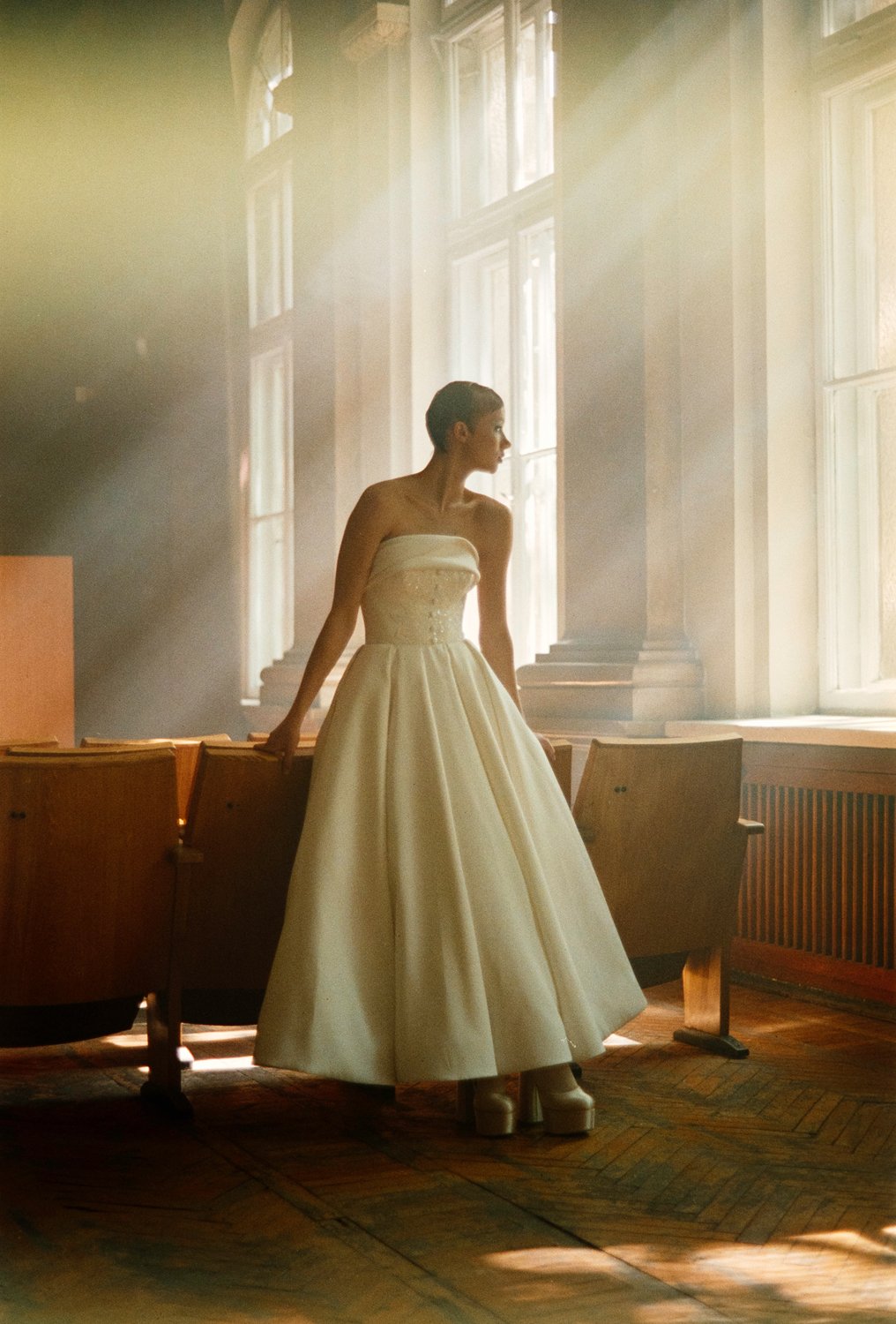 A woman in a strapless cream bride's dress with a full skirt and platform shoes standing in a sunlit room with large windows and wooden chairs.