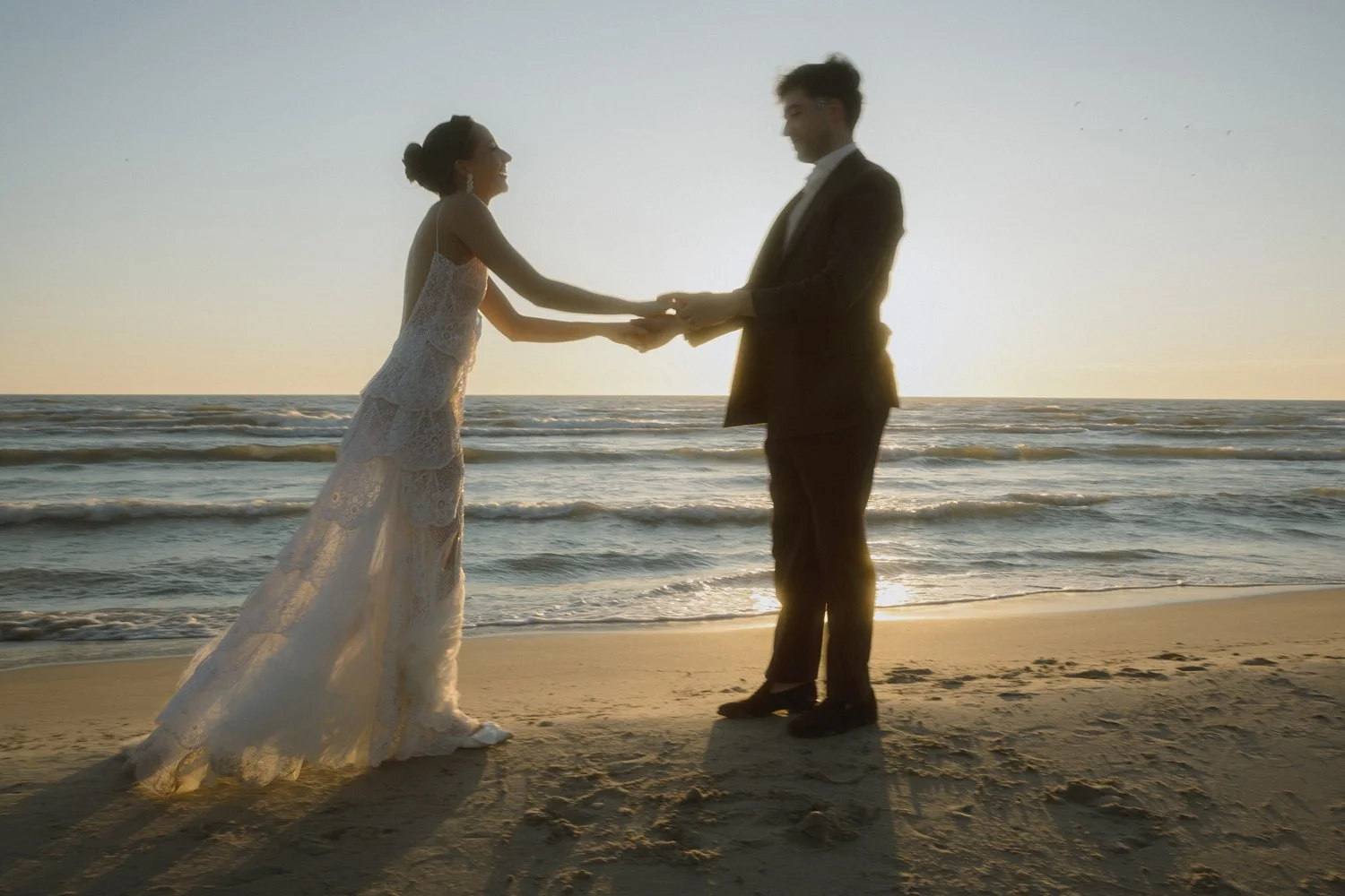 A bride and groom holding hands on a beach during sunset.