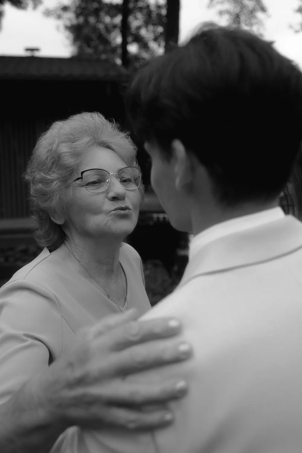 An elderly woman with glasses and curly hair is facing a younger man; they are holding each other in a close embrace in an outdoor setting.