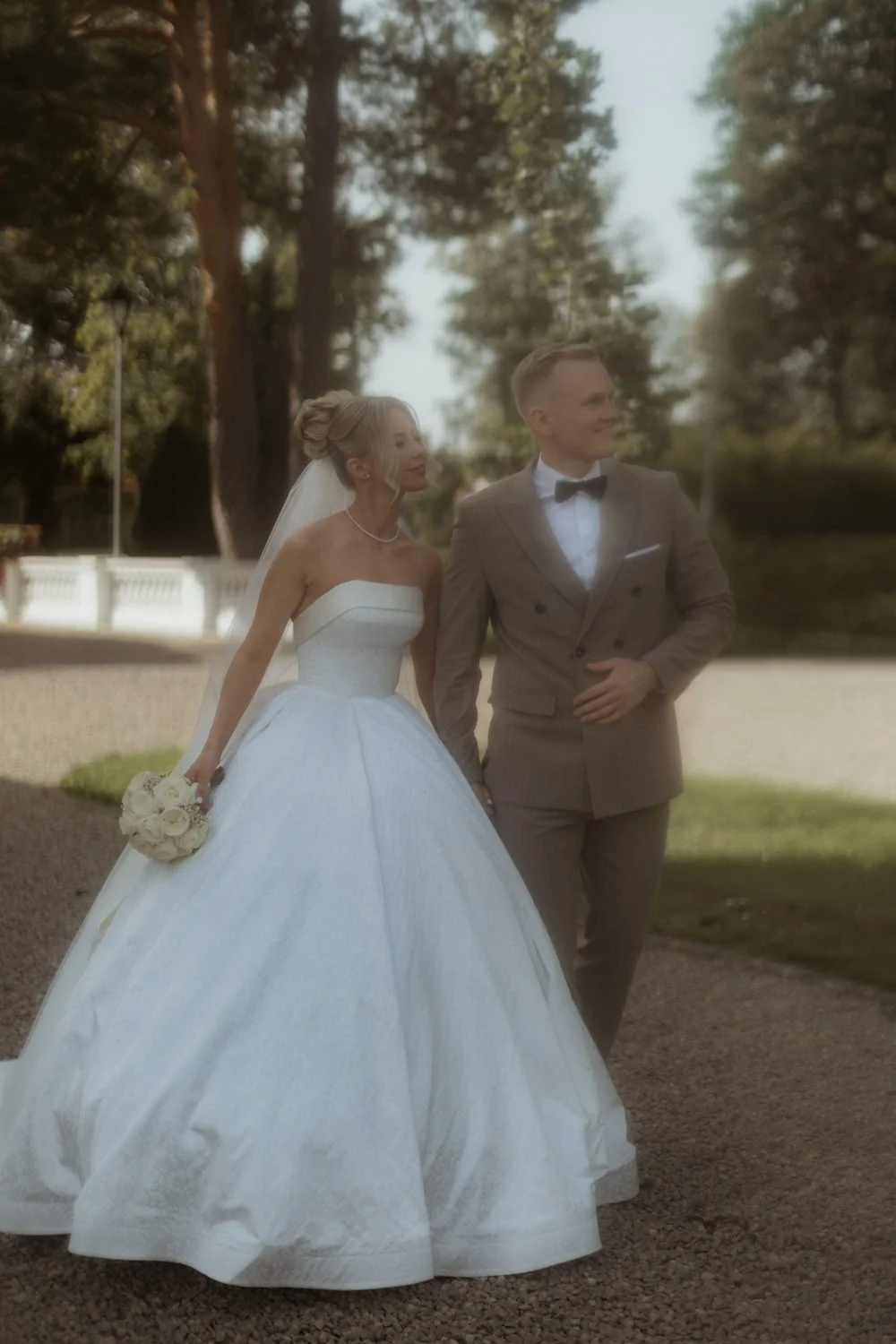 A bride and groom walking outdoors in a park setting, with the bride holding a bouquet of white roses, and both dressed in formal wedding attire.