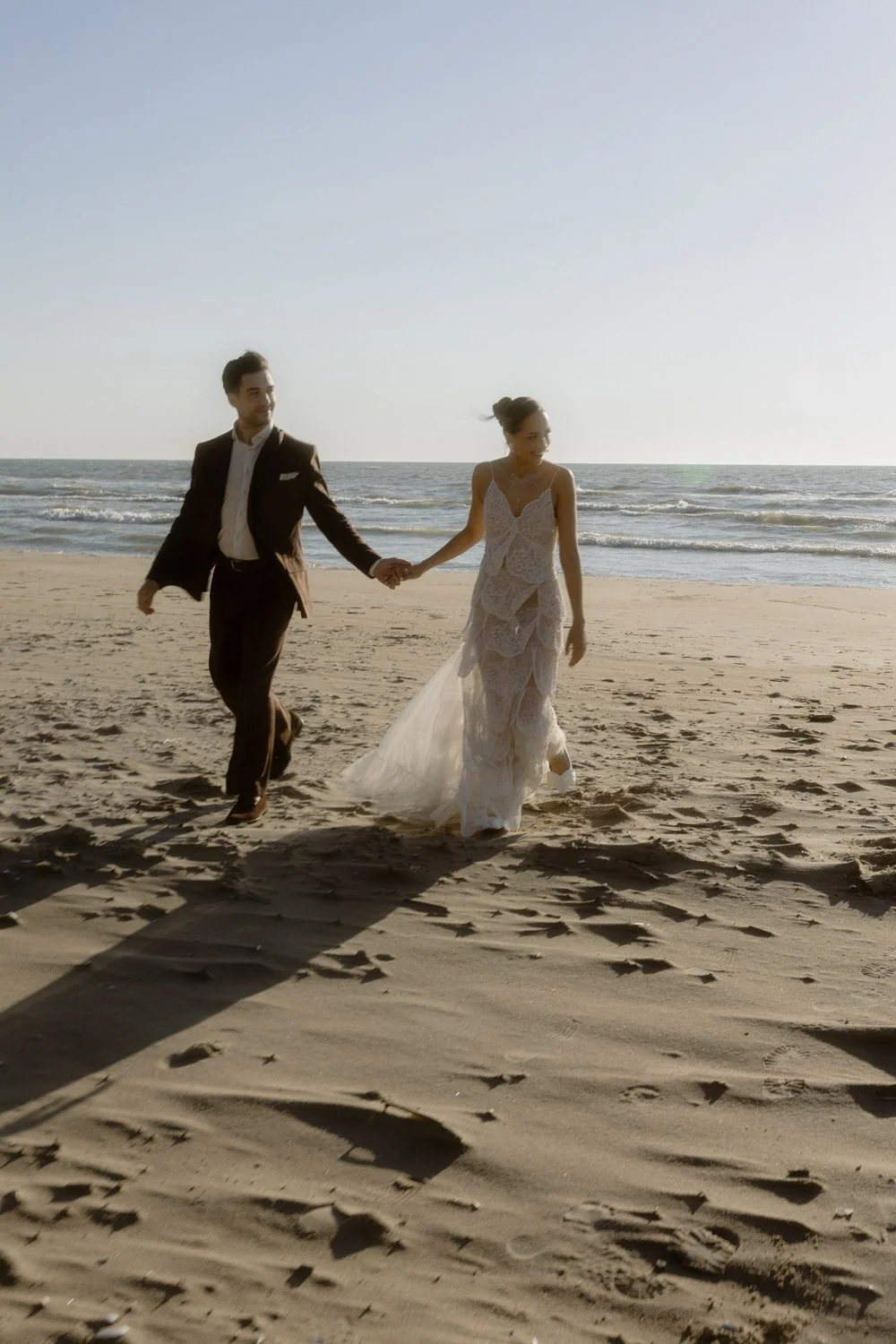 A couple dressed in wedding attire walking hand in hand on a sandy beach with the ocean in the background during sunset.