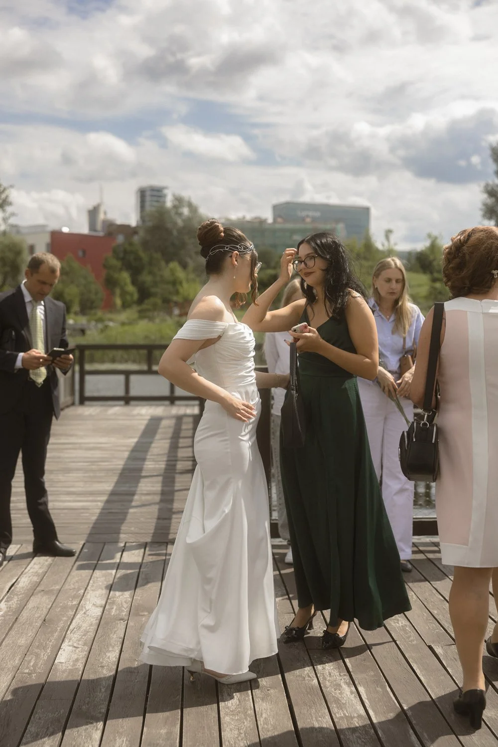 A woman in a white wedding dress and a woman in a dark green dress are talking and laughing on a wooden deck outdoors with other women and a man in a suit nearby, under a partly cloudy sky.
