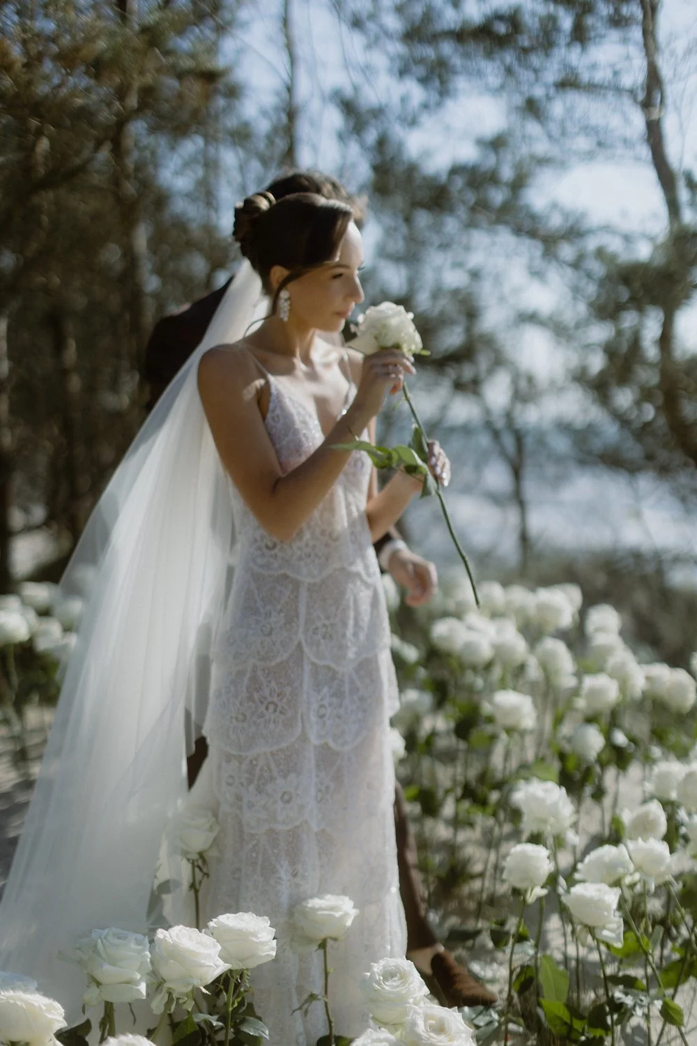 A bride in a lace wedding dress holding a white rose, standing among white roses outdoors with trees in the background.