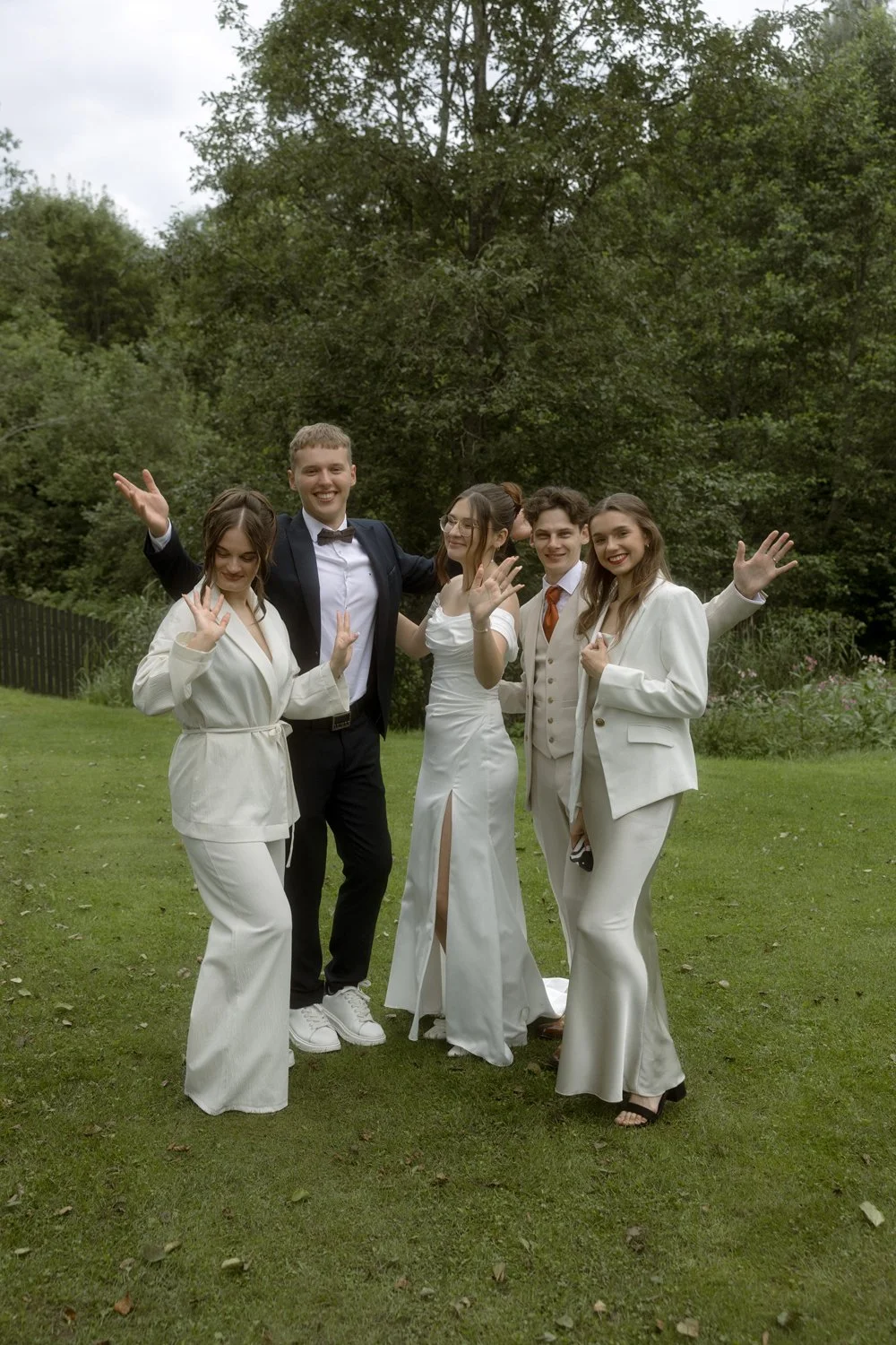A group of five young adults dressed in formal attire standing on a grassy field outdoors, smiling and waving at the camera.