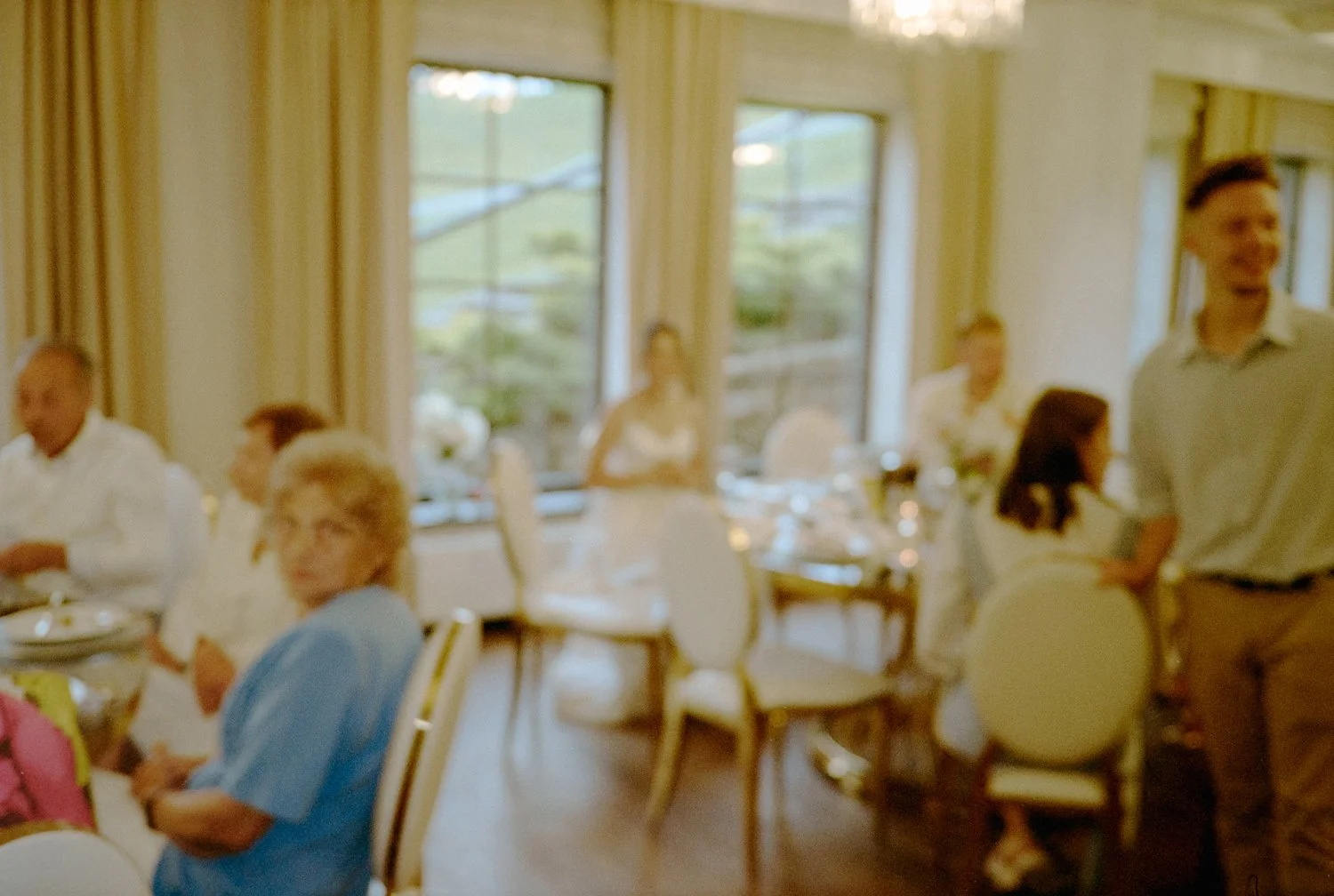A group of people gathered in a dining room with large windows and cream-colored chairs, some seated and some standing, with a woman in a white dress in the background.