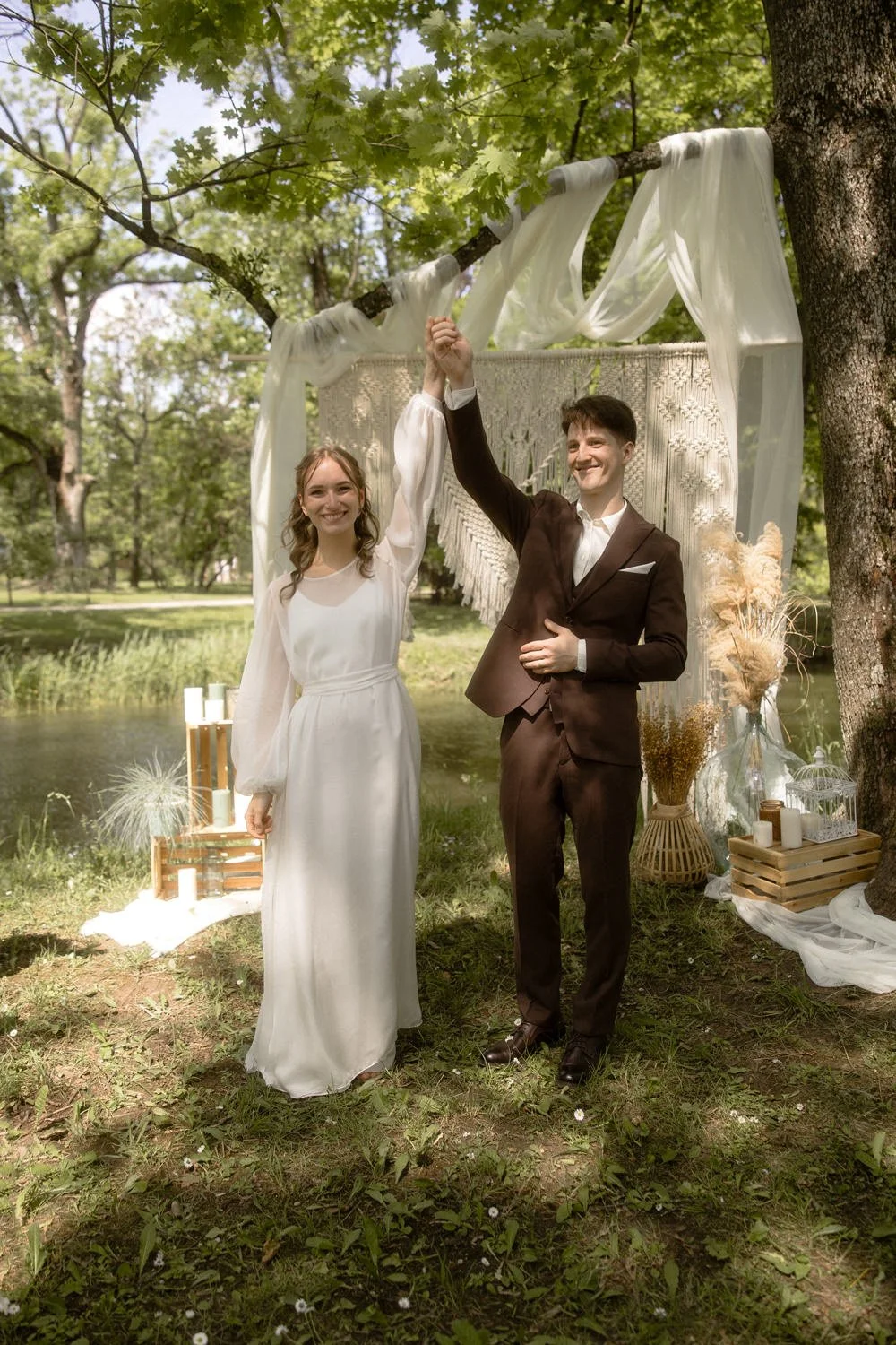 A bride and groom celebrating their wedding outdoors under a decorated tree, holding hands up in a victory pose.