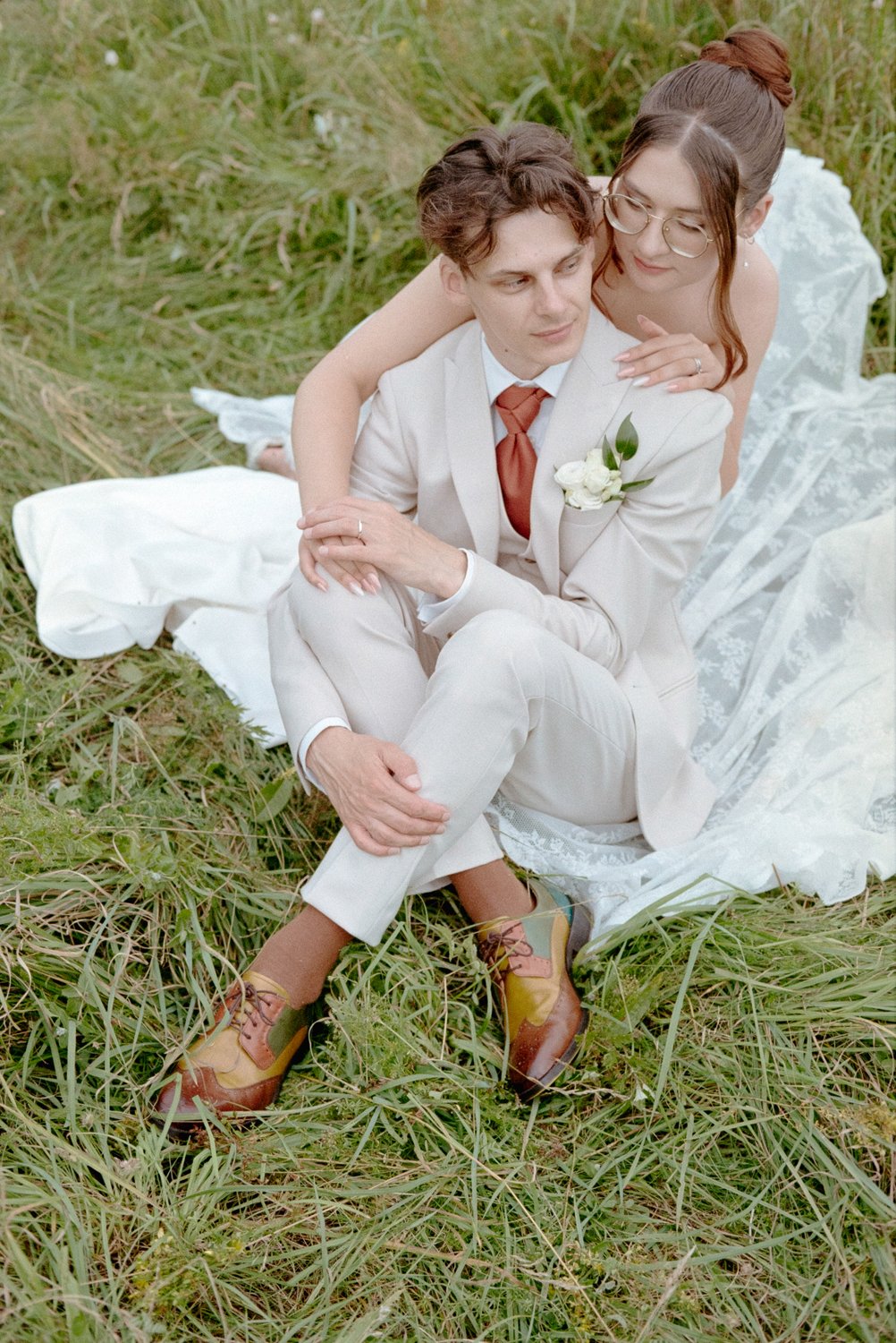 A couple in wedding attire sitting on grass, the woman embracing the man from behind, outdoors with green grass and some white fabric nearby.