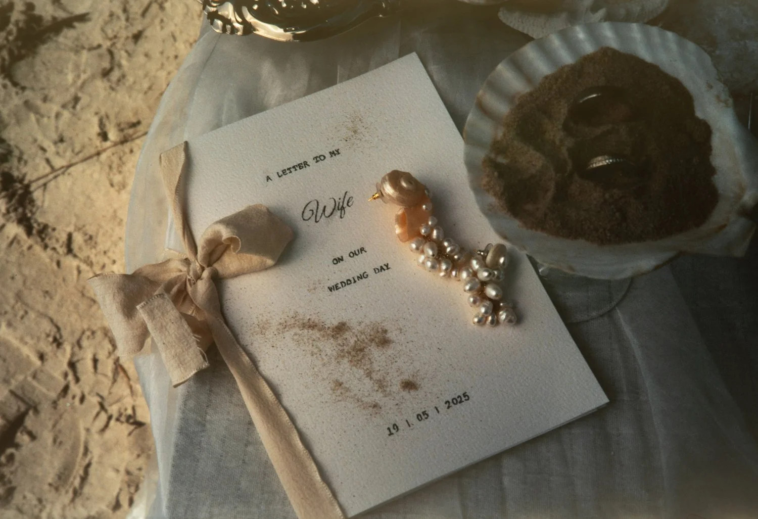 A white card with black text reading 'A letter to my wife on our wedding day,' dated May 19, 2025. The card is decorated with a beige ribbon, a pearl necklace, and a pearl earring. There is a small bowl of coffee with foam and some cocoa powder on to