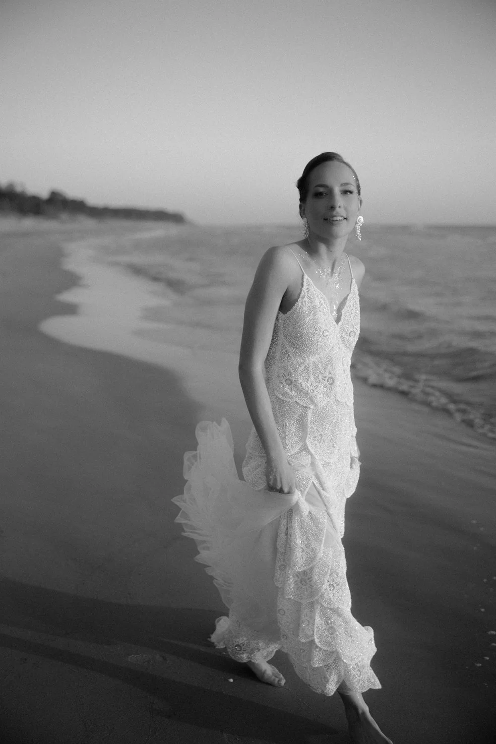 A woman in a wedding dress walking along the beach during sunset or dusk, holding the back of her dress, with waves in the background.
