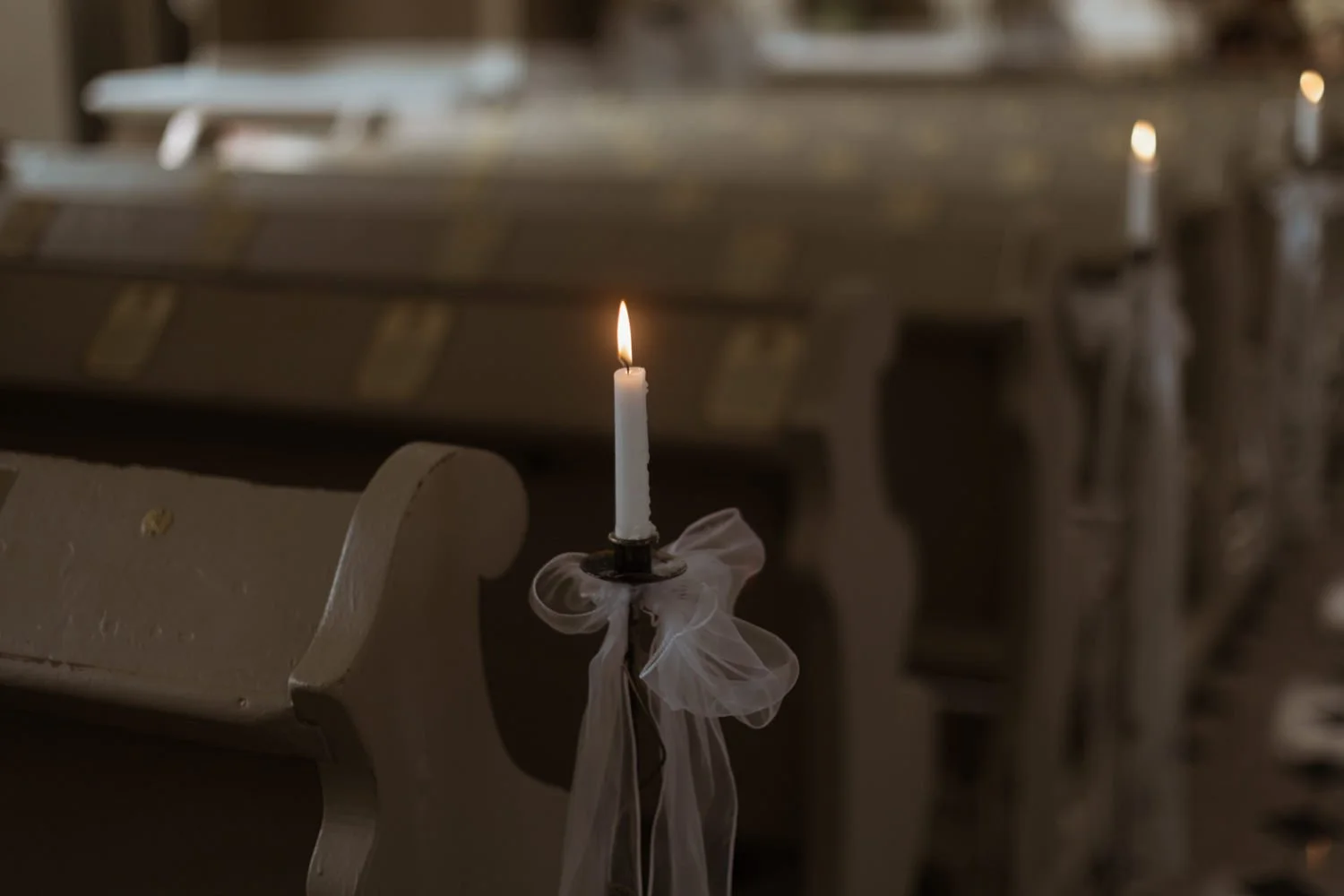 A lit white candle with a ribbon tied around its base, placed on a candle holder attached to the side of a pew in a church, with a row of similar candles and pews in the background.