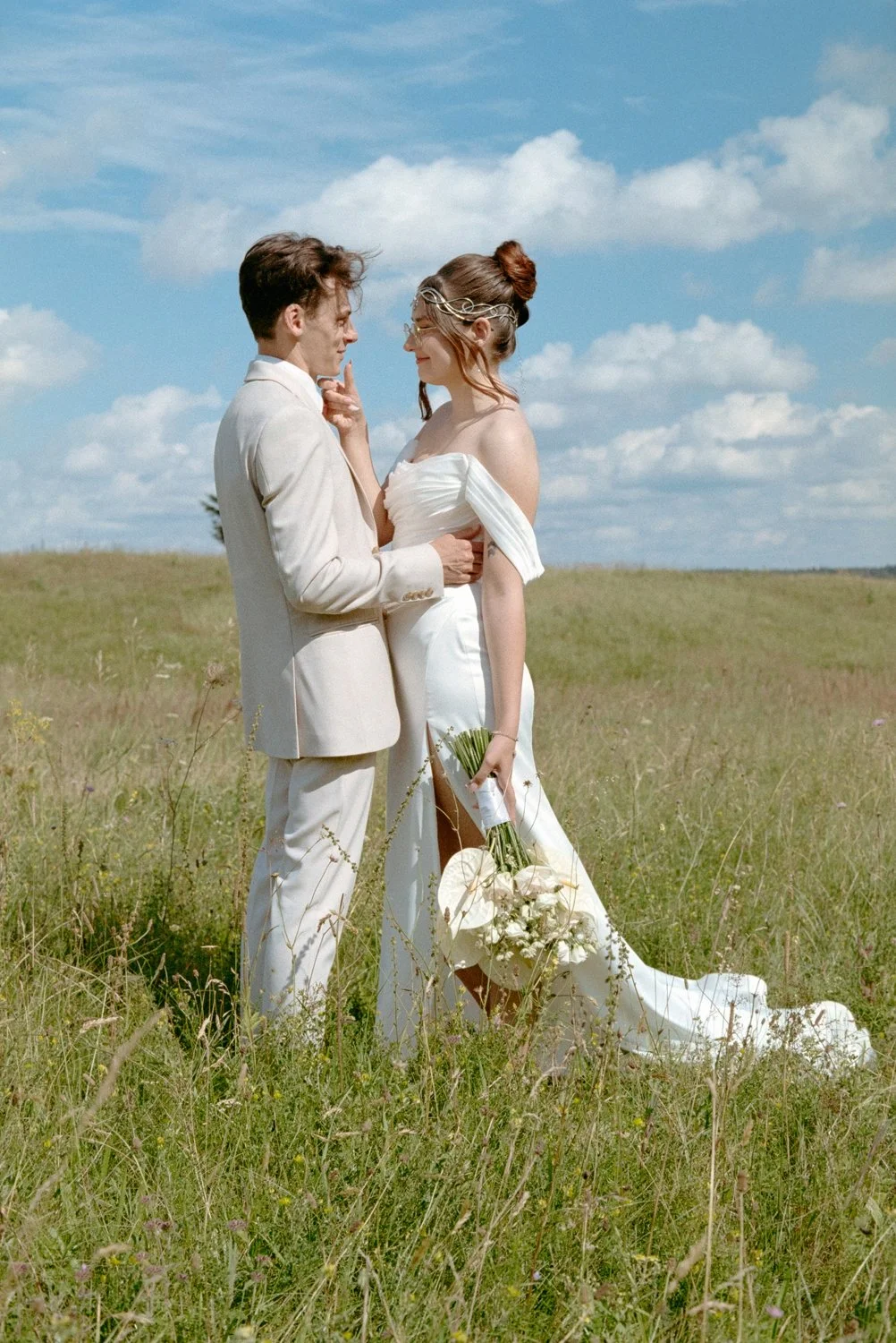 A couple in wedding attire standing in a grassy field on a sunny day with a blue sky and clouds, exchanging a tender moment.