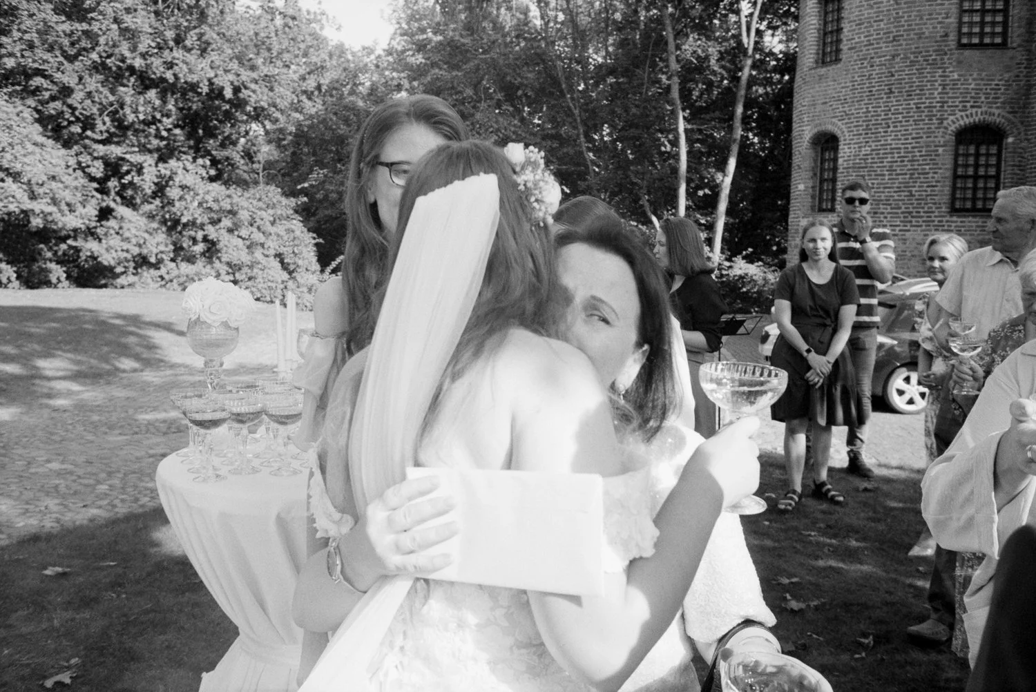 A black and white photograph of a wedding celebration outdoors, showing two women hugging and smiling, with a group of guests in the background and a brick building.
