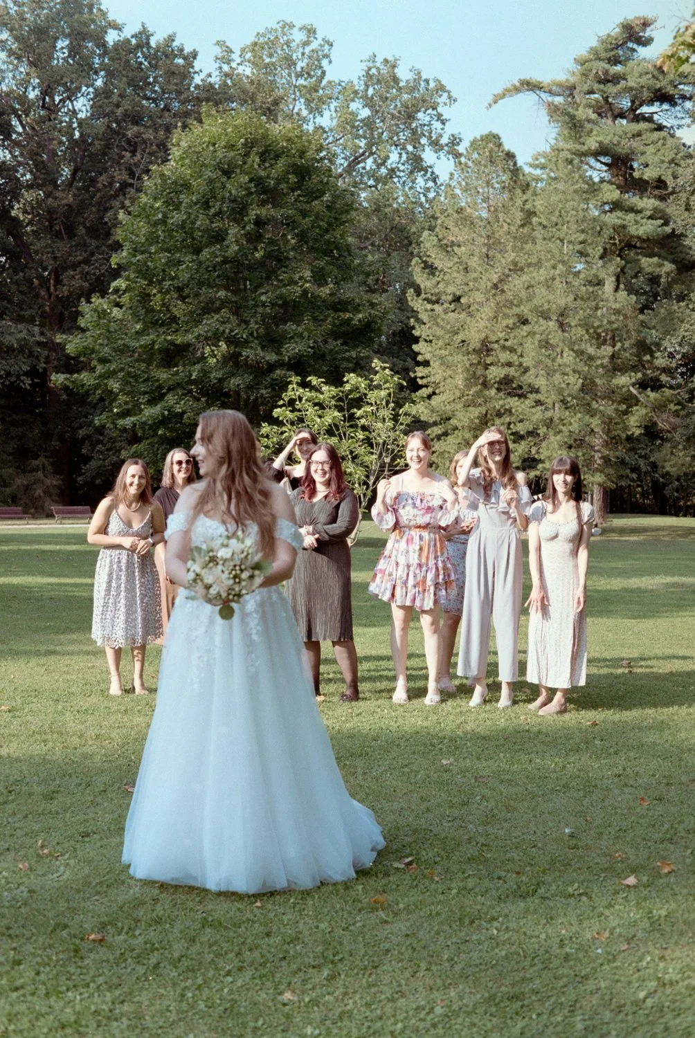 A bride in a white wedding dress holding a bouquet faces away from the camera. She is looking at seven women standing in a line behind her on a grassy field, with trees and a clear blue sky in the background. The women are dressed in summer dresses a