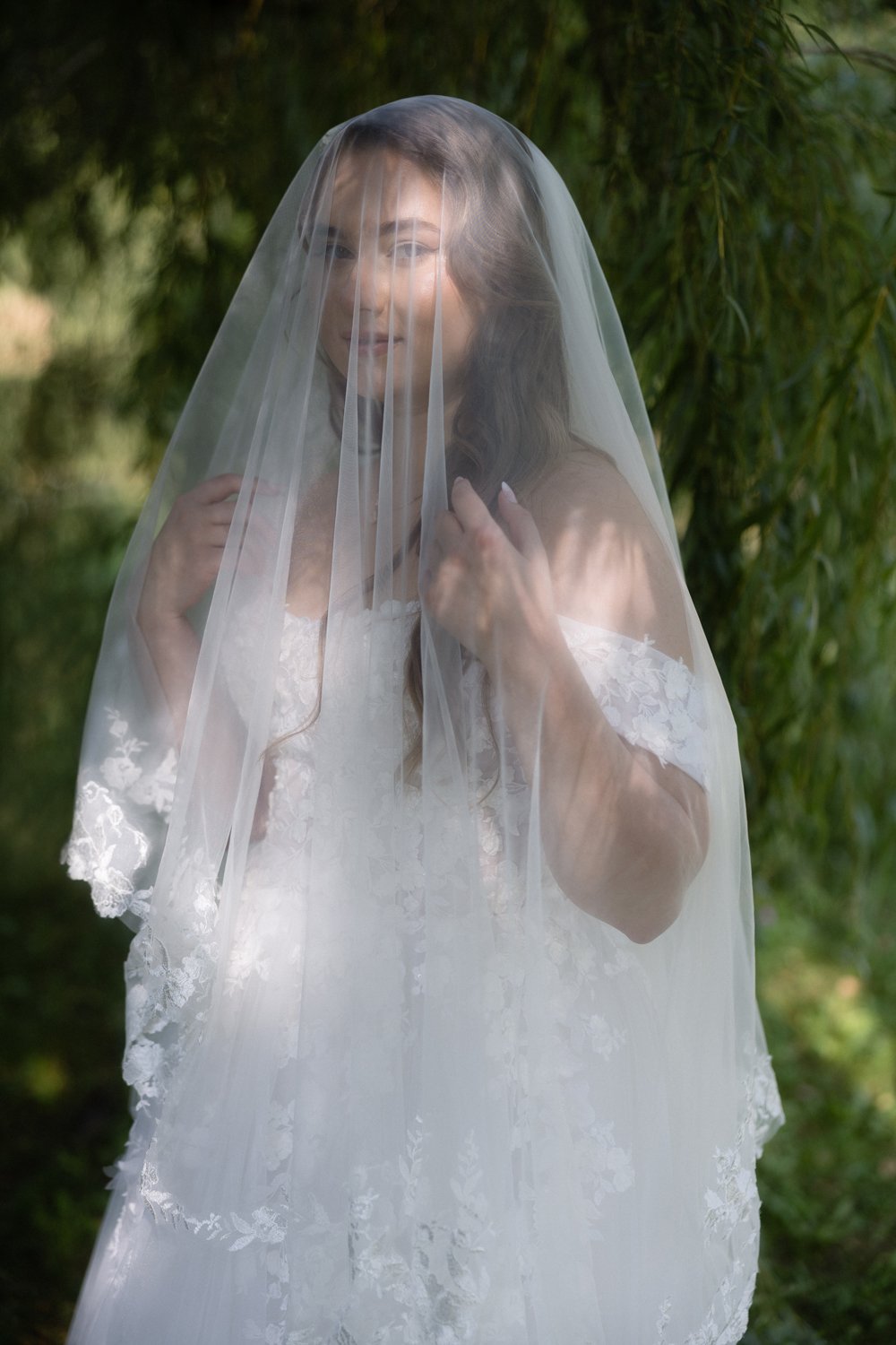 A woman in a white lace dress, wearing a veil, standing outdoors with greenery in the background, smiling.