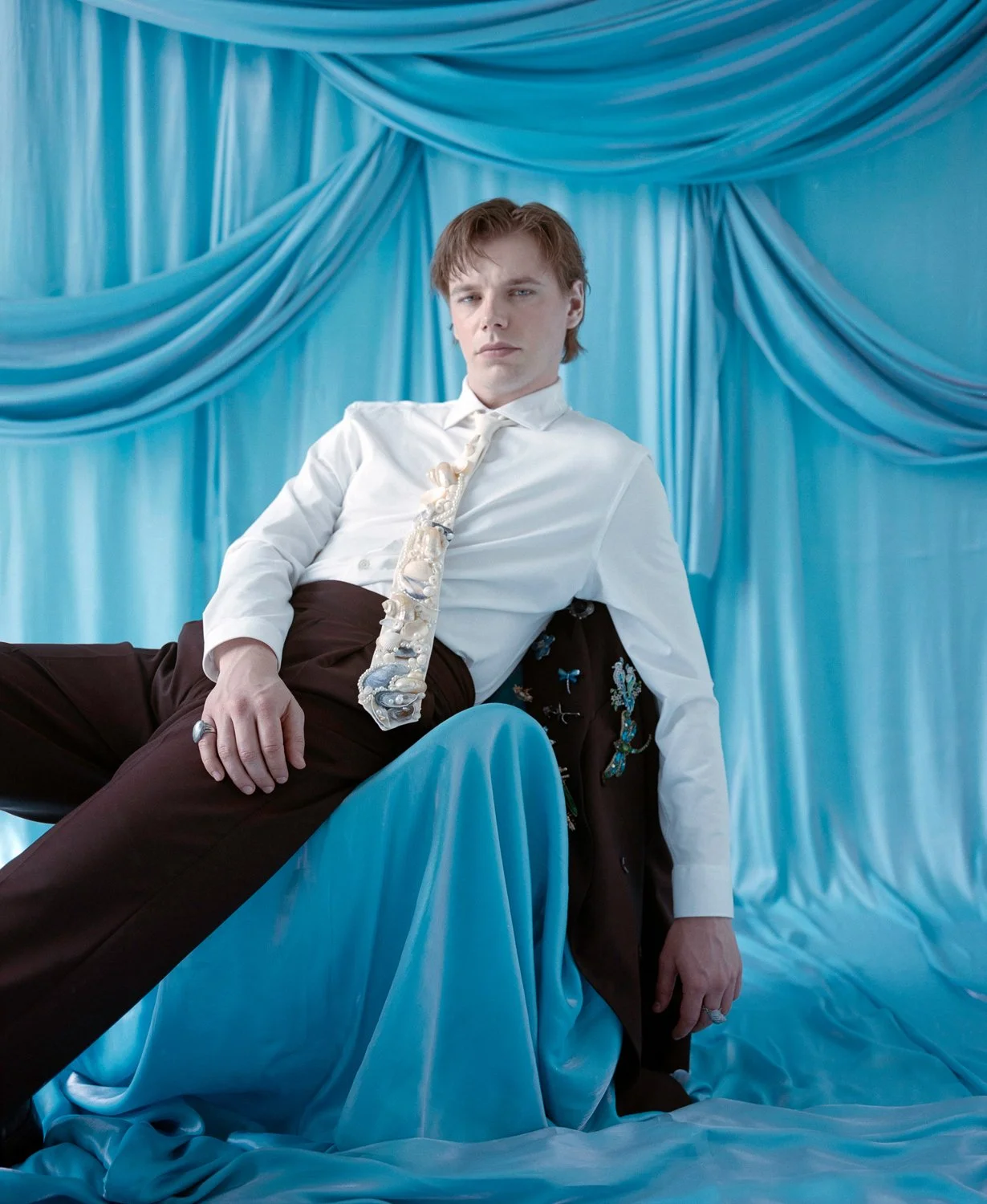 A young man with light brown hair, wearing a white shirt, tie, and dark brown pants, reclining against a chair with a blue silky fabric backdrop. The setting has an elegant, vintage vibe.