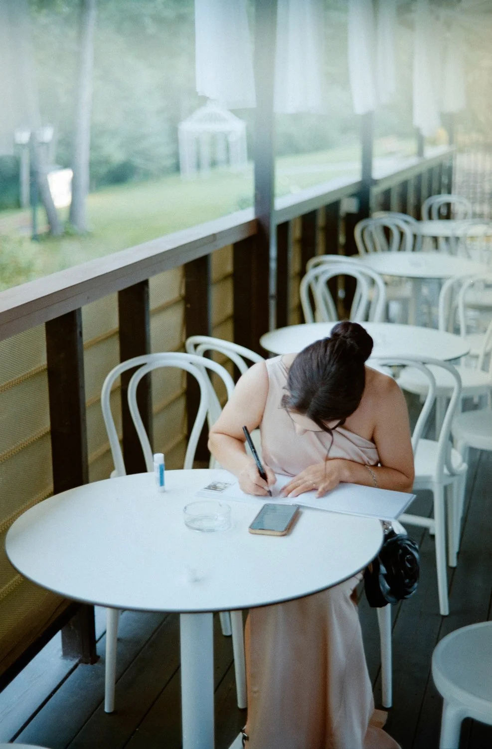 A woman sitting at an outdoor table on a porch, writing in a notebook, with a smartphone, a small glue stick, and a glass of water on the table. There are empty white chairs around her and a screened-in railing.