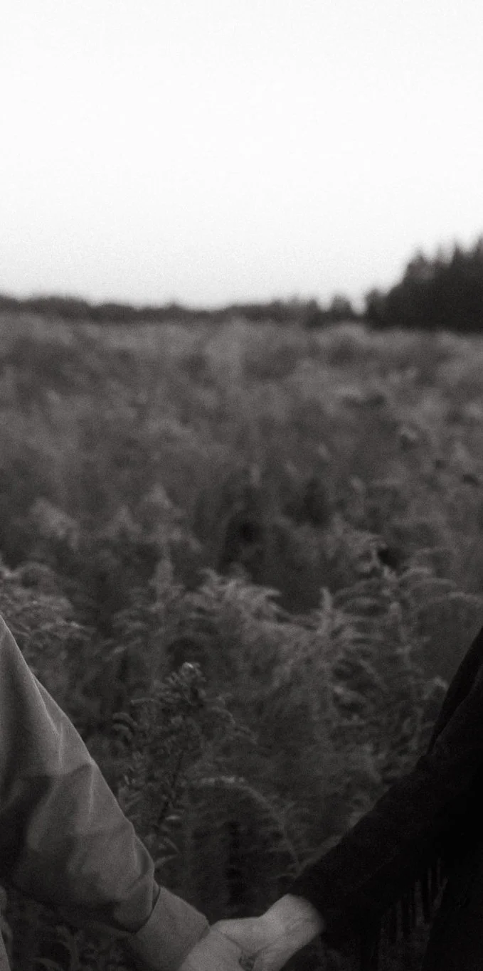 Black and white photo of two people holding hands in an outdoor natural setting with trees and plants in the background.