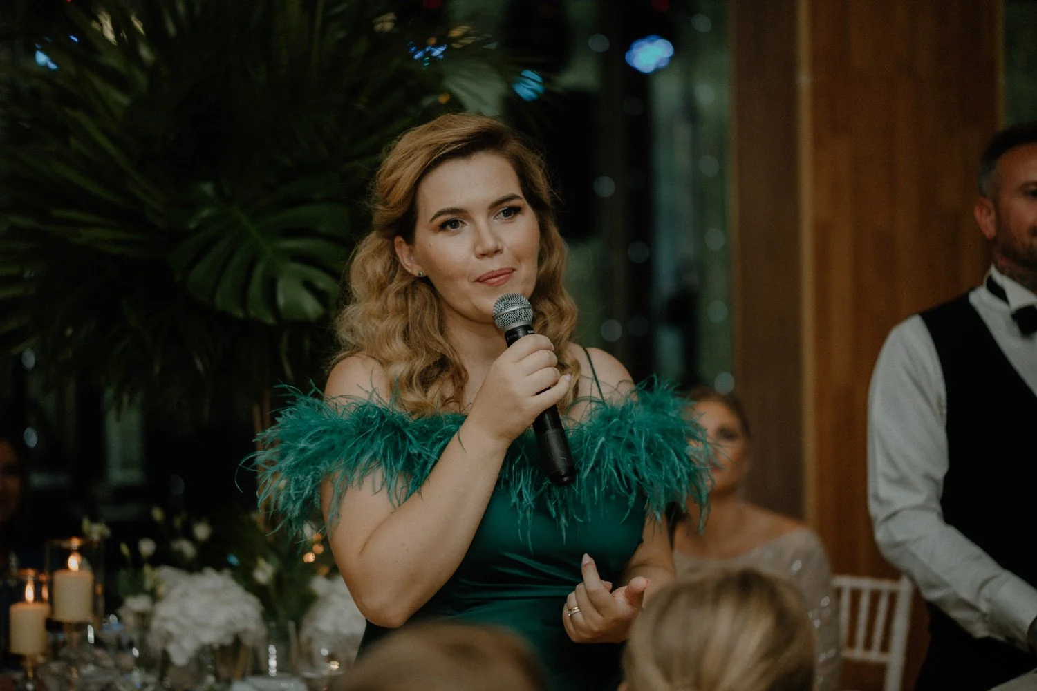 A woman in a green dress with feathered shoulders holding a microphone and speaking at an indoor event, with people seated at tables and large green plants in the background.