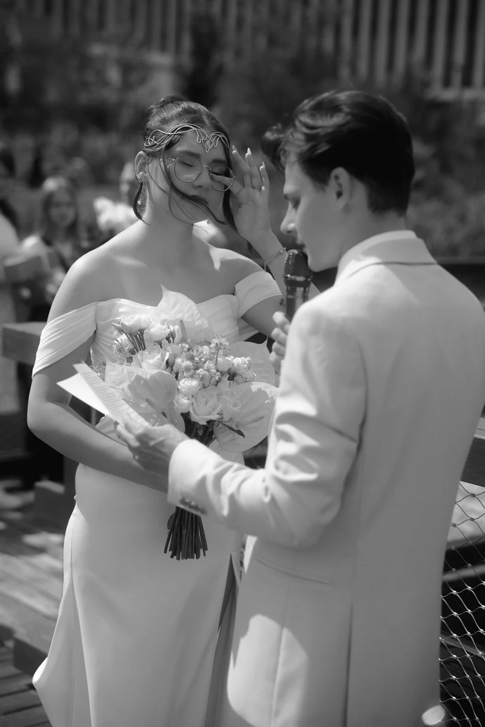 A black-and-white photo of a wedding ceremony showing a bride, wearing glasses and a white dress, holding a bouquet, with her eyes closed, as the groom, in a white suit, holds a microphone and reads from a paper, touching her forehead gently.